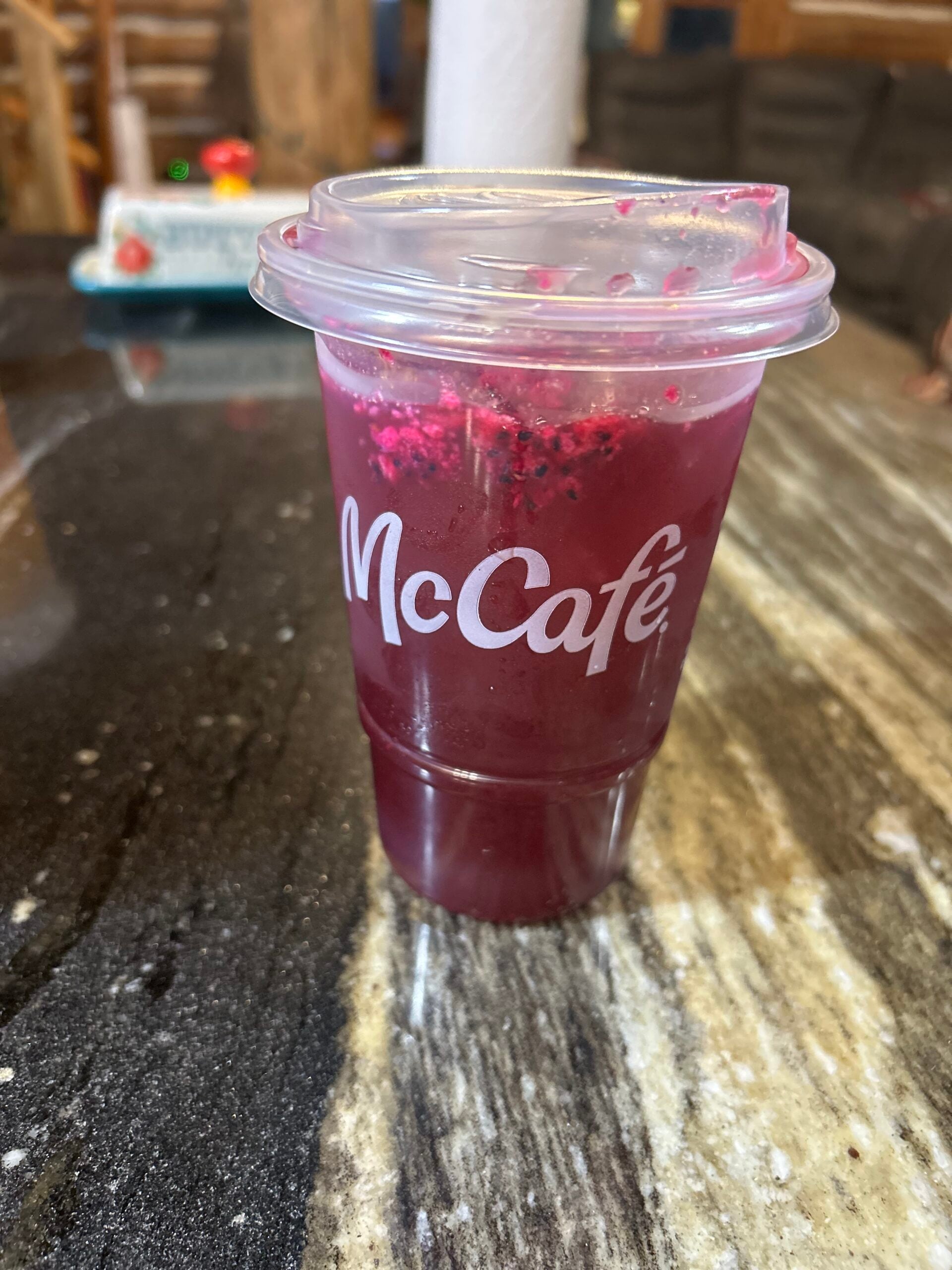 A clear McCafé cup filled with a vibrant purple drink sits on a dark marble countertop, with a plastic lid on top. The background is blurred, showing a cozy indoor setting.