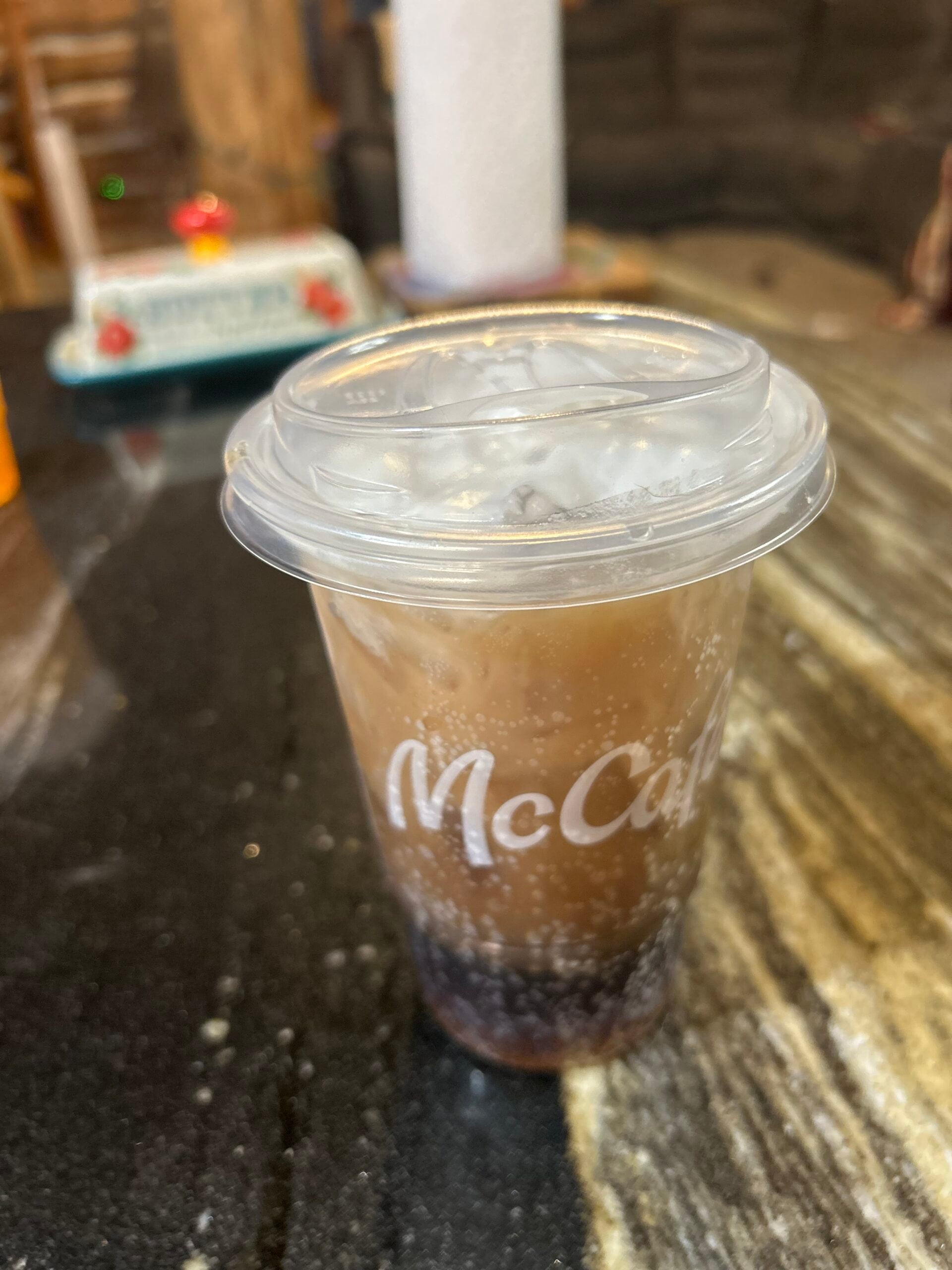 A clear plastic McCafé cup with a lid contains a layered iced drink, dark at the bottom and creamy brown on top, sitting on a marbled black and beige countertop.