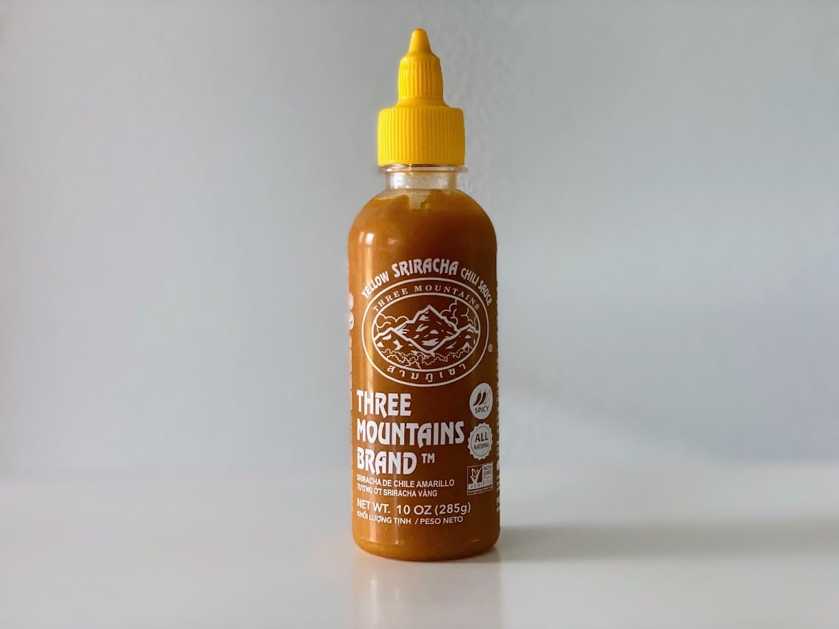 A bottle of Three Mountains Brand Sriracha chili sauce with a yellow cap and white text on the label, standing on a light surface against a plain background.