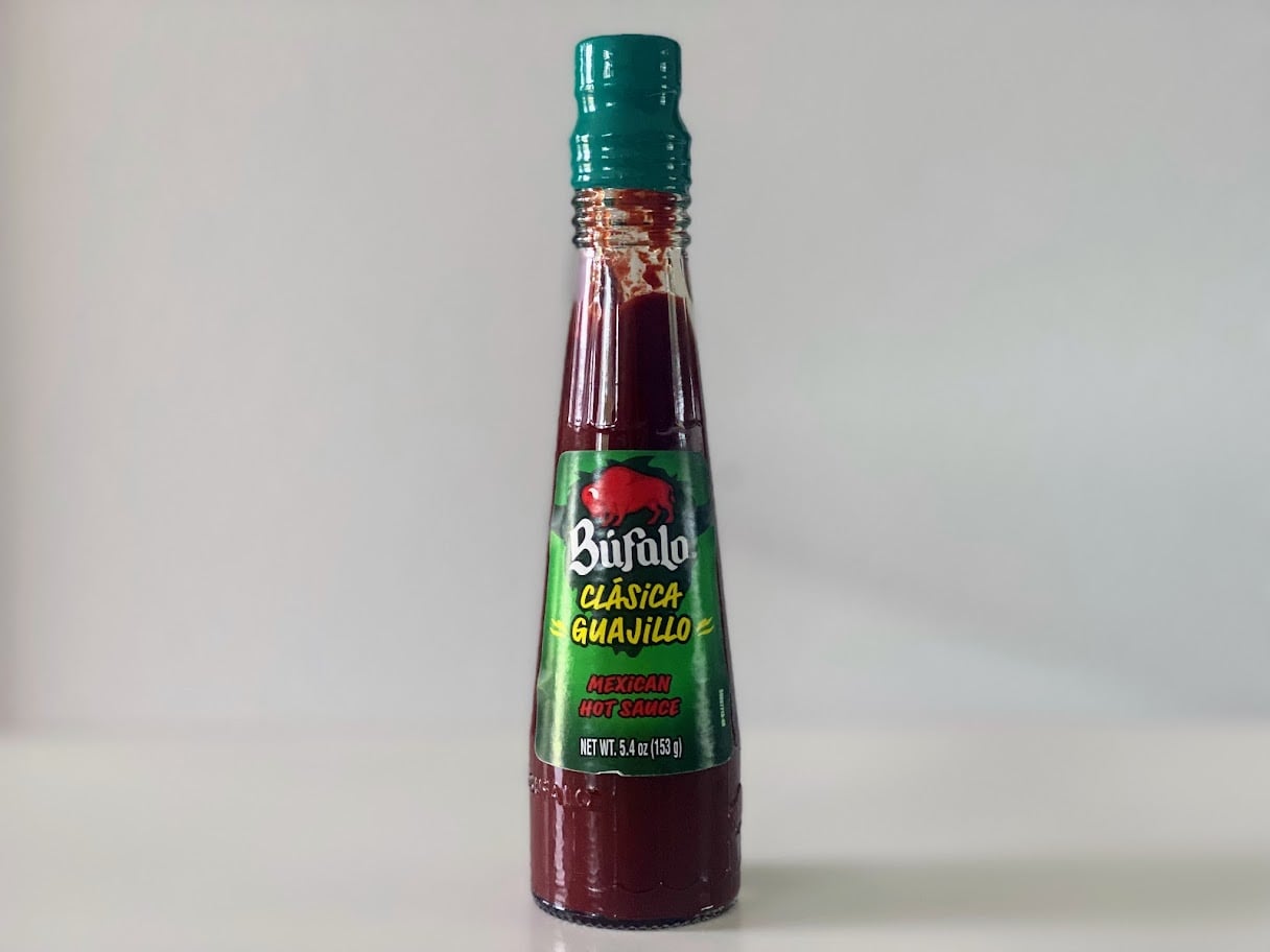 A bottle of Bufalo Clasica Guajillo Mexican hot sauce with green and red labeling sits on a white surface against a light gray background.