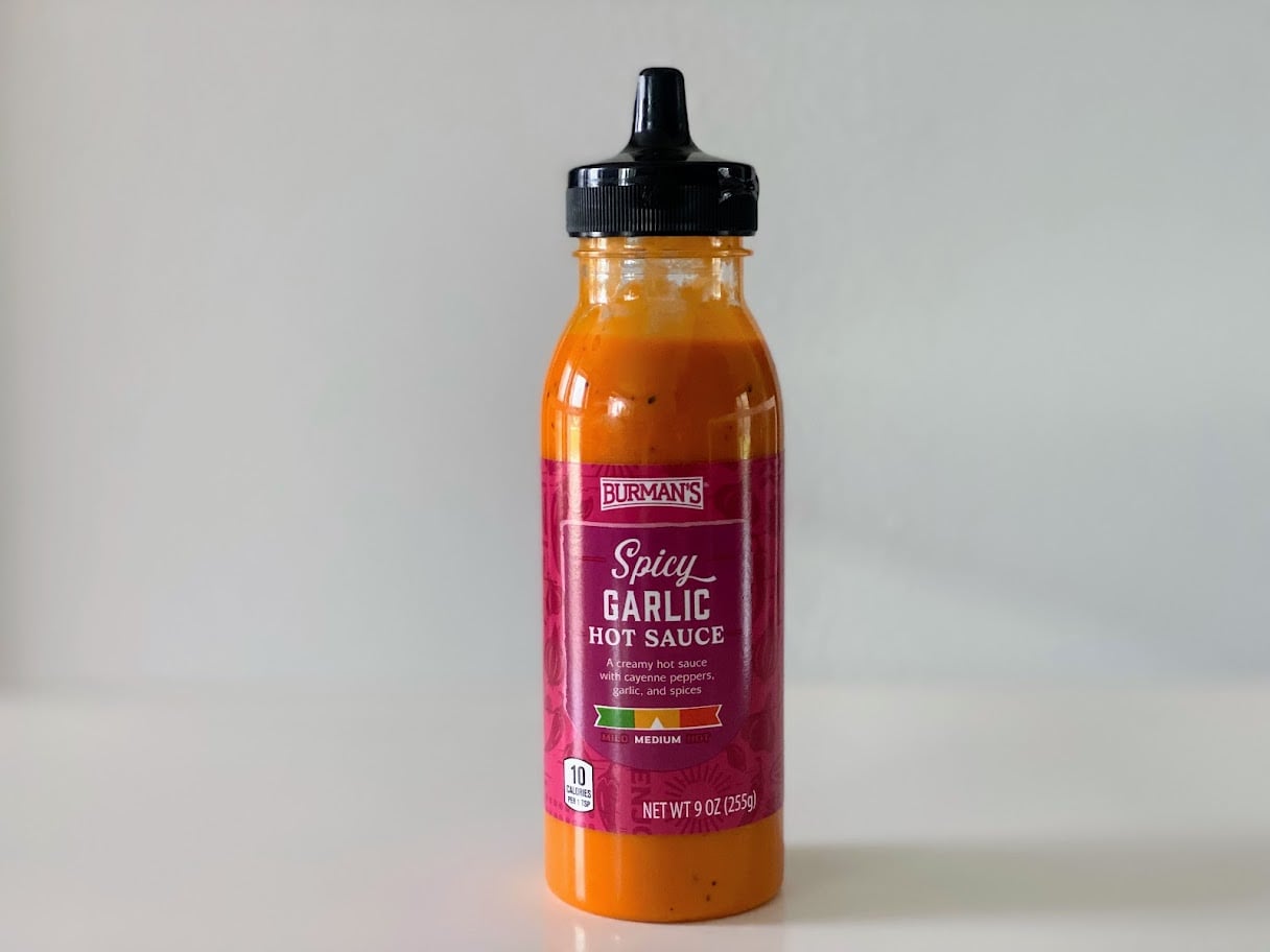 A bottle of Burman's Spicy Garlic Hot Sauce with a black cap sits on a white surface against a light gray background. The label is red and states the sauce is medium heat and 9 ounces.