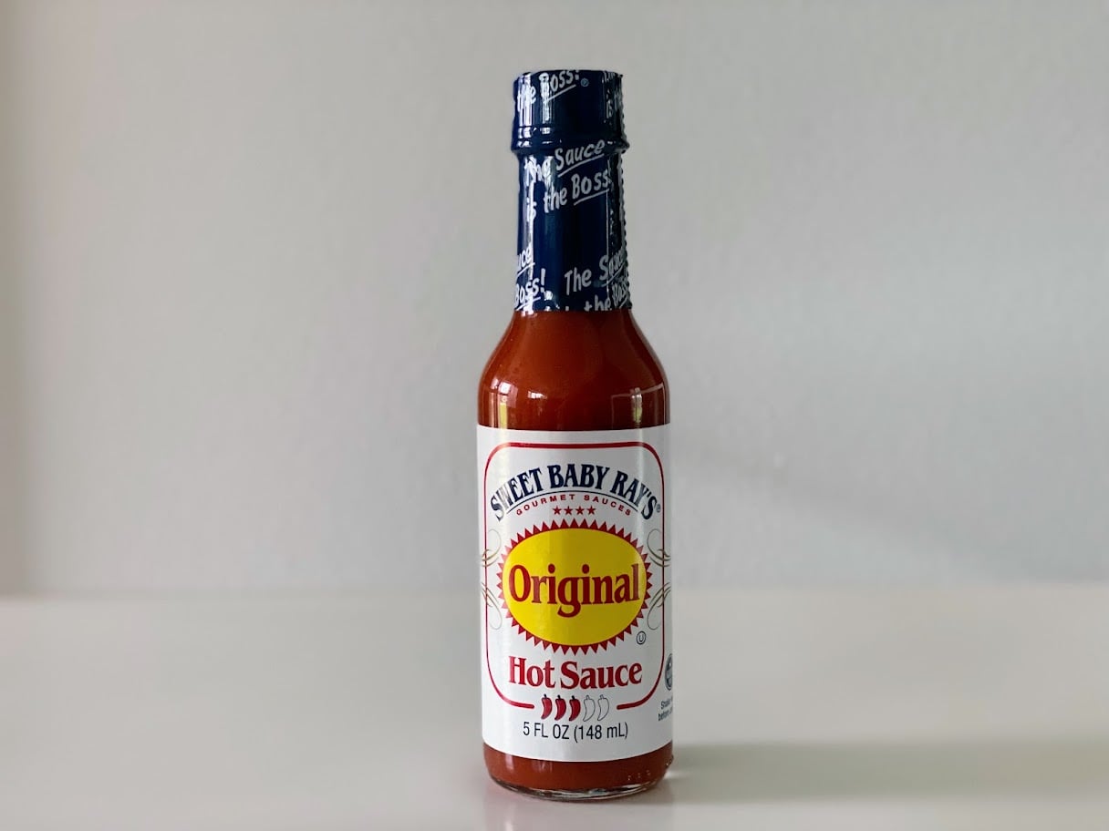 A 5 fl oz bottle of Sweet Baby Ray’s Original Hot Sauce sits on a white surface with a plain light gray background. The bottle has a blue cap and a white label with red and yellow accents.