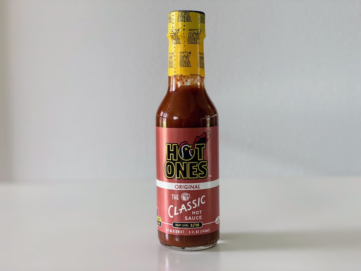 A bottle of Hot Ones Original "The Classic" hot sauce stands on a white surface, with a red label and yellow cap featuring the Hot Ones logo.