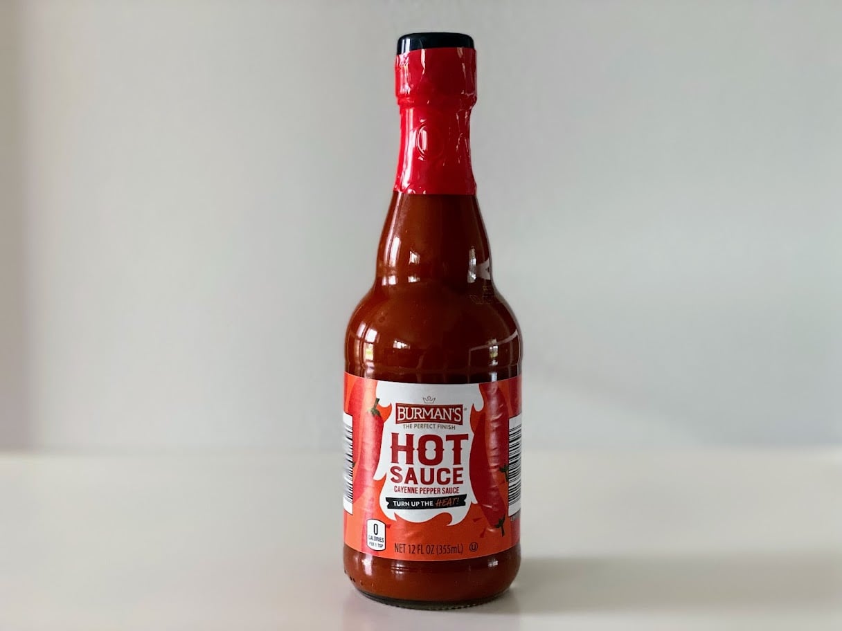 A bottle of Burman's Hot Sauce with a red cap and label stands on a white surface against a plain light background. The label reads "Gluten Free" and "Cayenne Pepper Sauce.