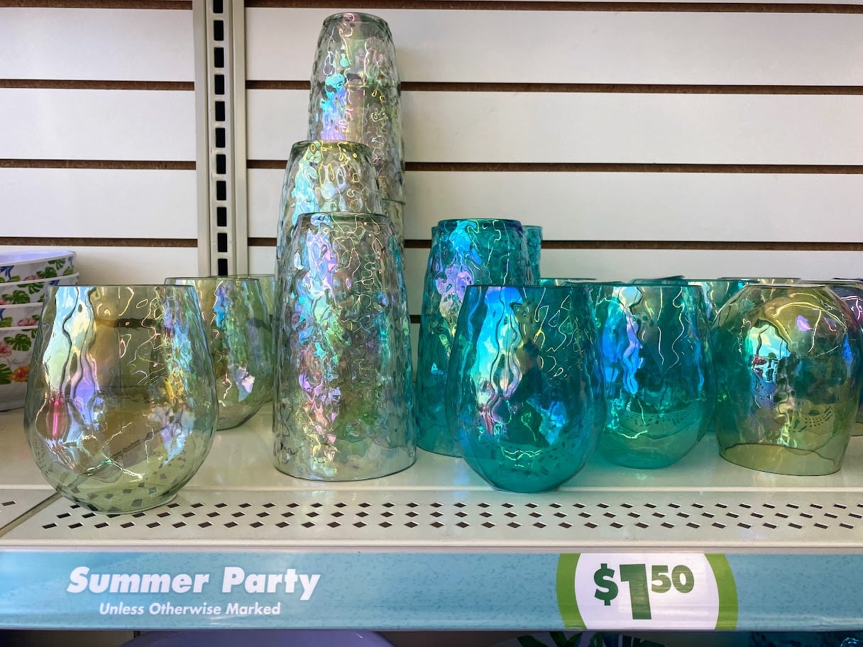 Iridescent blue and green glass cups and vases are displayed on a store shelf with a sign reading “Summer Party” and a price of $1.50.