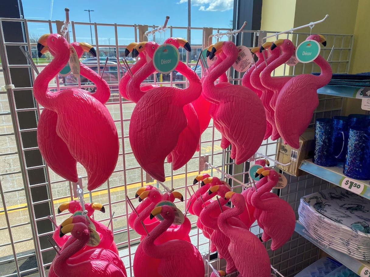 Bright pink plastic flamingo decorations hang on a metal display rack in a store, with sunlight streaming in from a window. Price tags are visible, and other colorful items are displayed nearby.