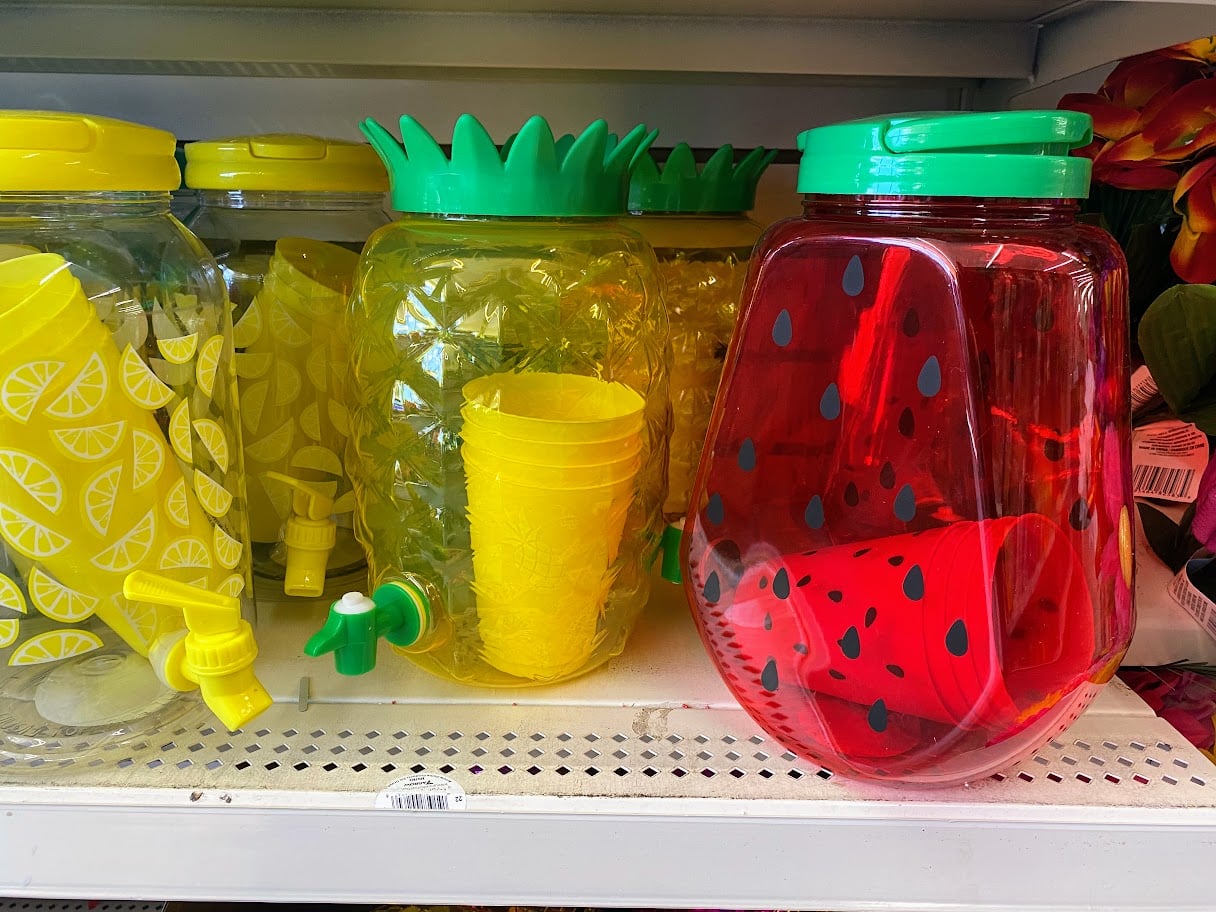 Plastic beverage dispensers and cups shaped like pineapples, lemons, and a red jar with black seeds resembling a watermelon are displayed on a store shelf.