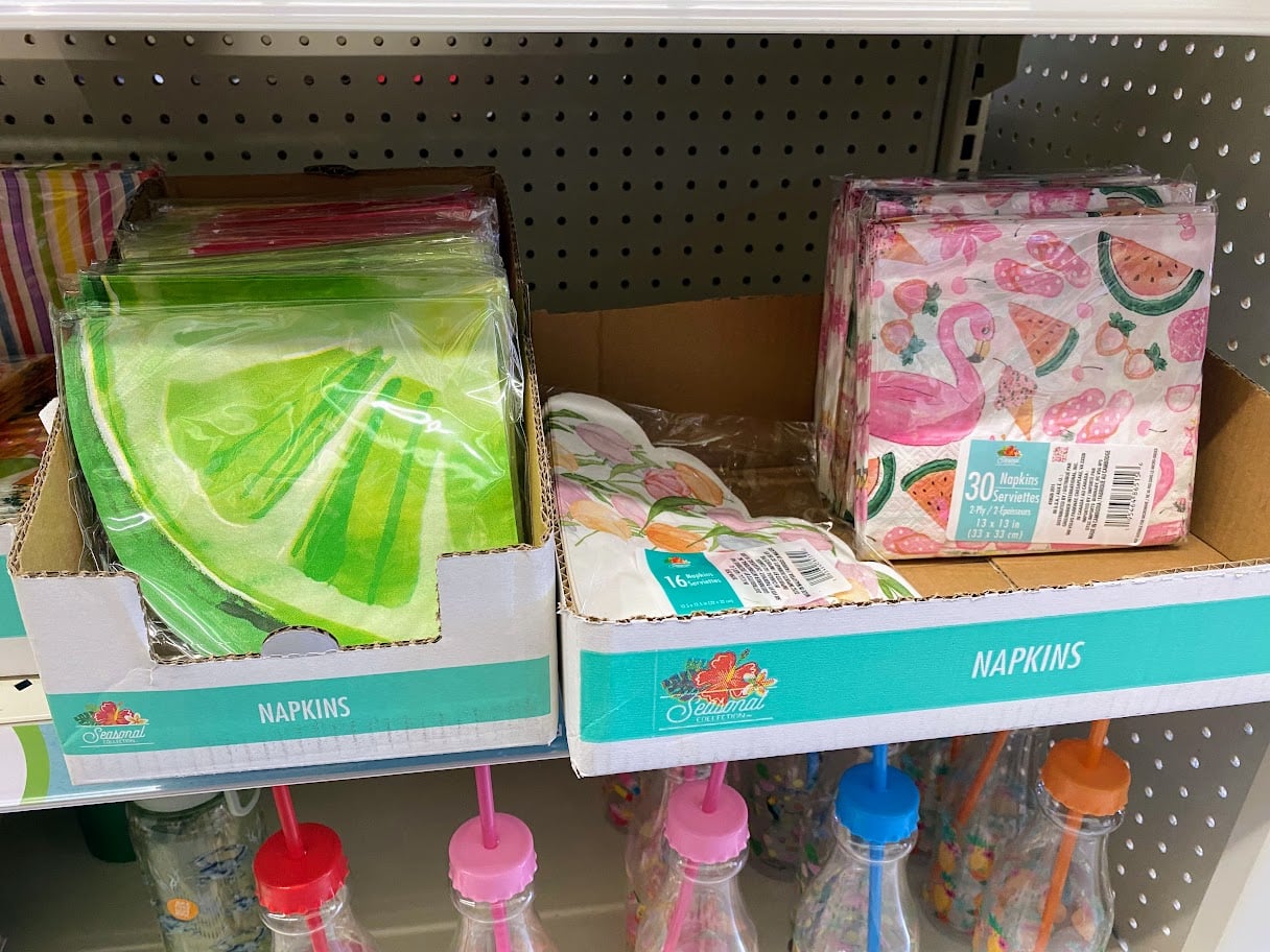 Two boxes of decorative napkins on a store shelf: one has green napkins shaped like watermelon slices, the other has pink napkins with flamingos, watermelons, and leaves. Colorful bottles are visible on the shelf below.