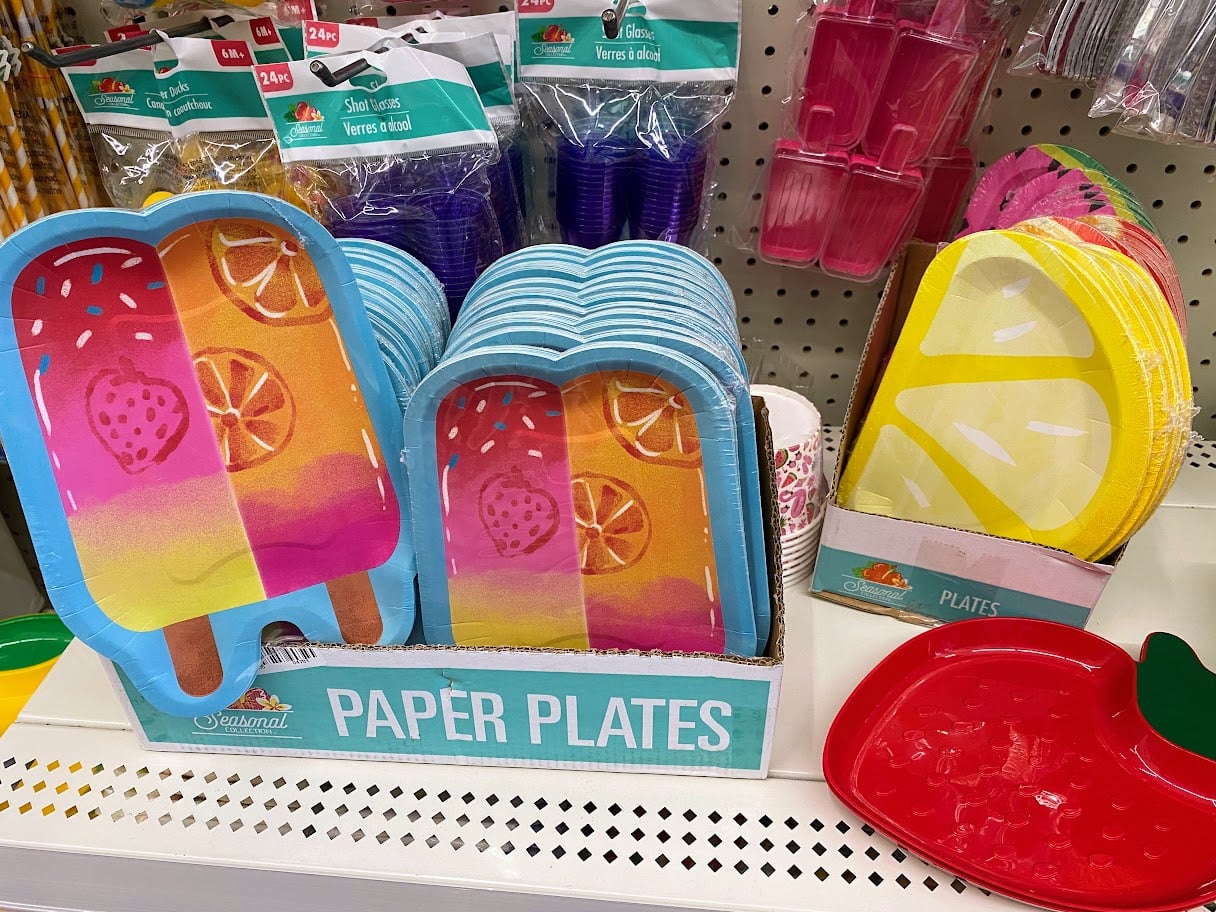 Colorful paper plates shaped like popsicles with fruit designs and lemon slices are displayed on a store shelf, along with shell-shaped purple shot glasses and pink trays in the background.