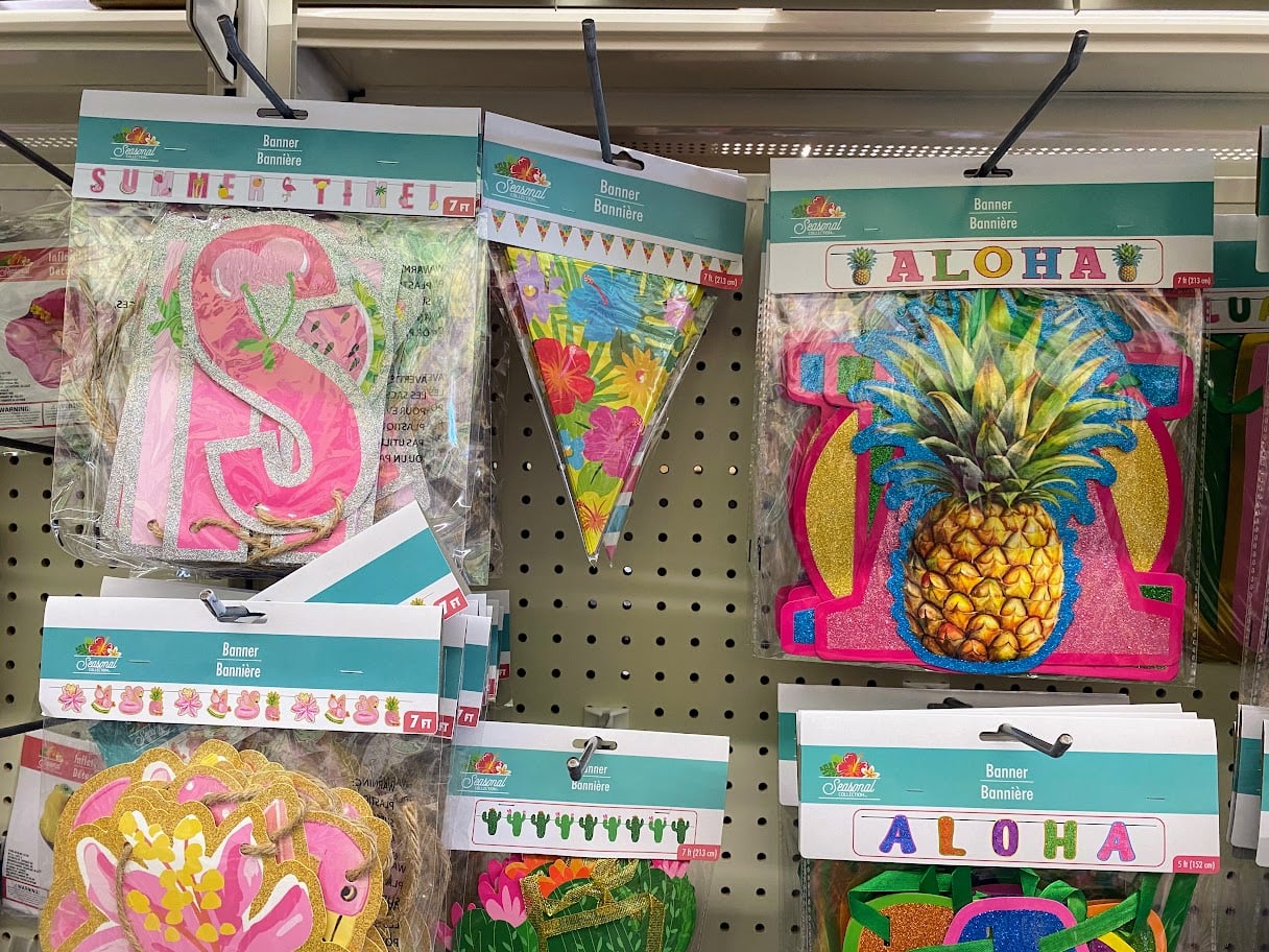 Colorful summer-themed banner decorations hang on a store display, featuring designs like pineapples, tropical flowers, and the words "SUMMER TIME" and "ALOHA" in vibrant colors.