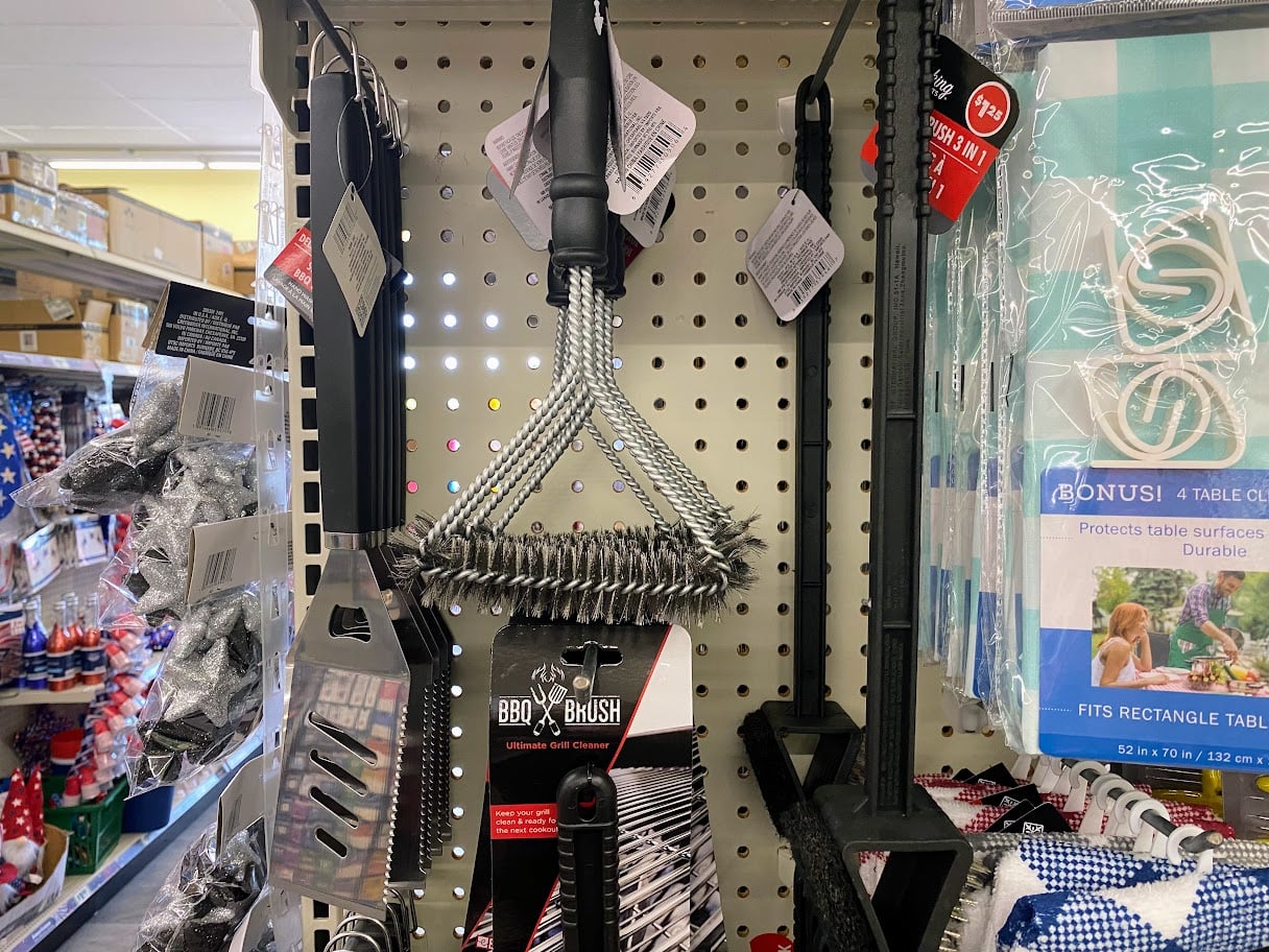 Various BBQ tools, including a grill brush and spatula, hang on a pegboard display in a store aisle. The items are packaged, and other kitchen and picnic supplies are visible nearby.