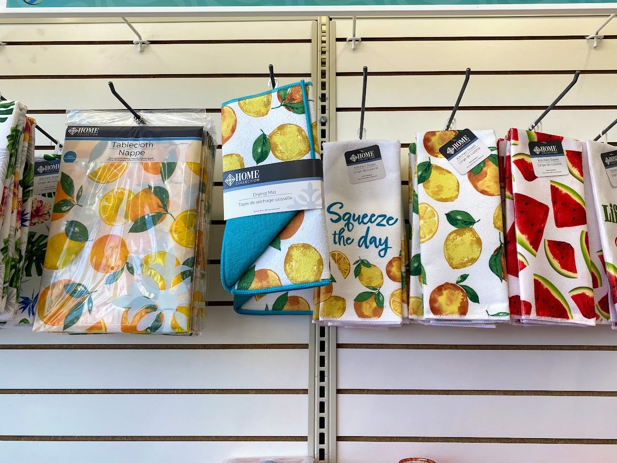 Colorful kitchen towels and napkins with fruit designs, including lemons, oranges, and watermelon, hang on display hooks against a white slat wall in a store. One towel says “Squeeze the day.”