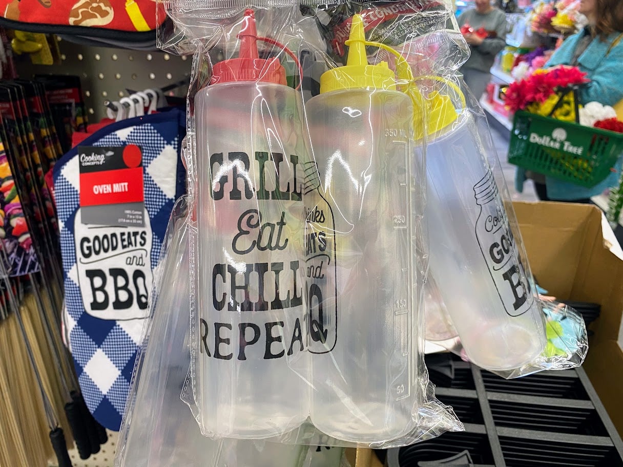 Clear plastic squeeze bottles with red and yellow caps hang in a store display. The bottles have text reading "Chill, Eat, Chill, Repeat." Nearby, blue oven mitts say "Good Eats and BBQ." Flowers are visible in the background.