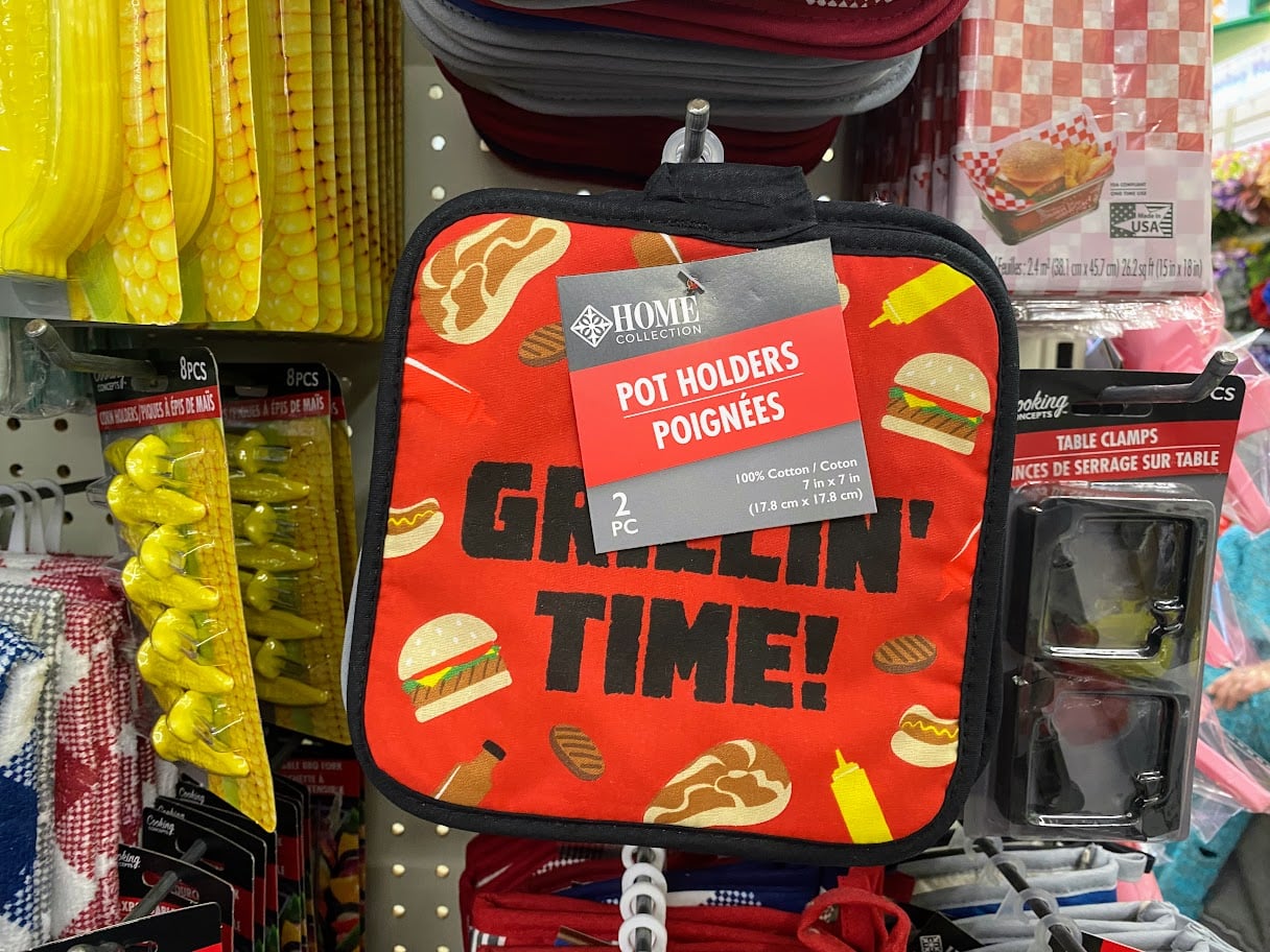 A red pot holder with images of burgers, hot dogs, and condiments hangs on a store shelf. The text on it reads "GRILLIN' TIME!" A label above says "Pot Holders" and nearby are other kitchen items.