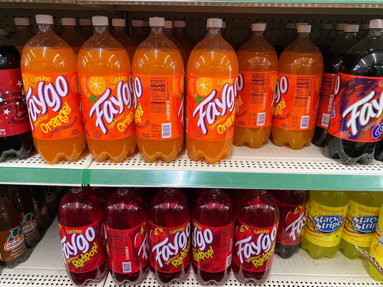 Bottles of Faygo soda in orange and redpop flavors are lined up on grocery store shelves, with other sodas like Starry Pineapple and Starry Stripe nearby.