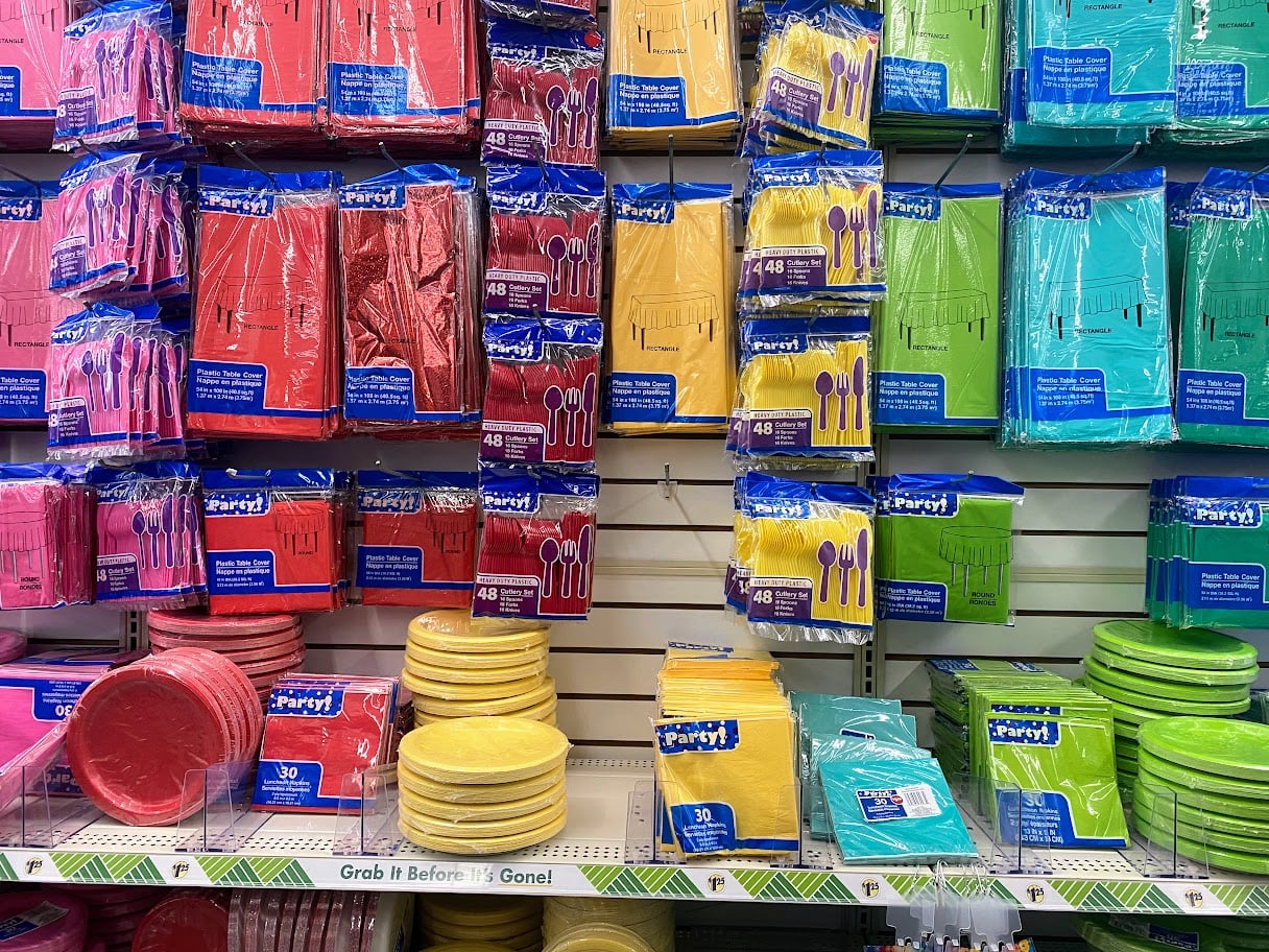 Colorful disposable party supplies, including plates, napkins, and utensils, are displayed on store shelves in red, pink, yellow, blue, green, and turquoise packaging. Items are neatly organized by color.