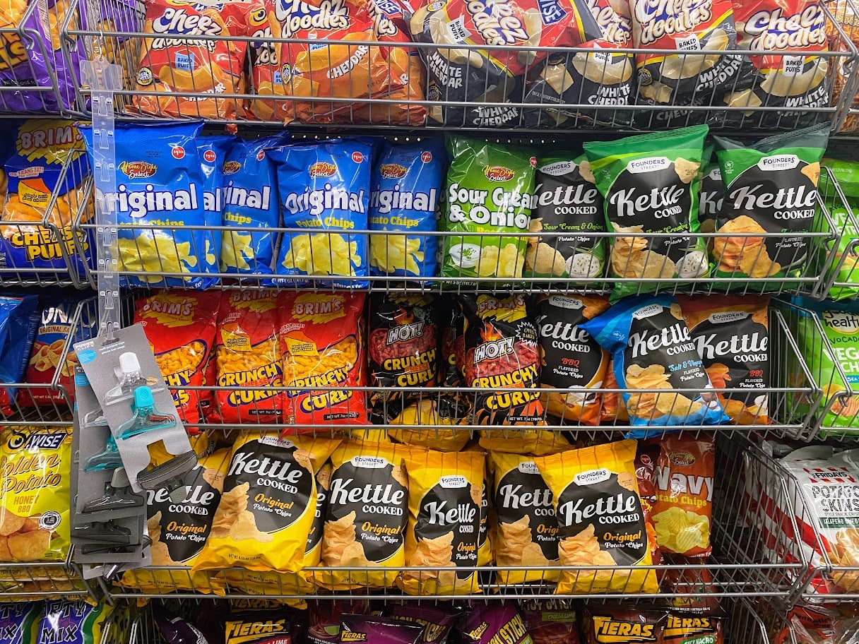 Bags of assorted chips, including potato chips, kettle chips, and cheese snacks, are arranged on metal shelves in a store. The shelves also display a few small non-food items on the left side.