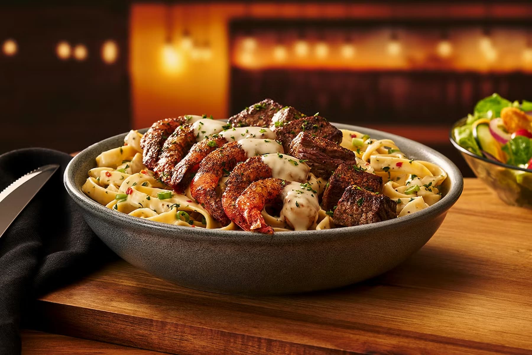 A bowl of fettuccine pasta topped with grilled steak, shrimp, and creamy sauce, garnished with herbs, sits on a wooden table next to a knife, black napkin, and a side salad. Warm, softly-lit background.