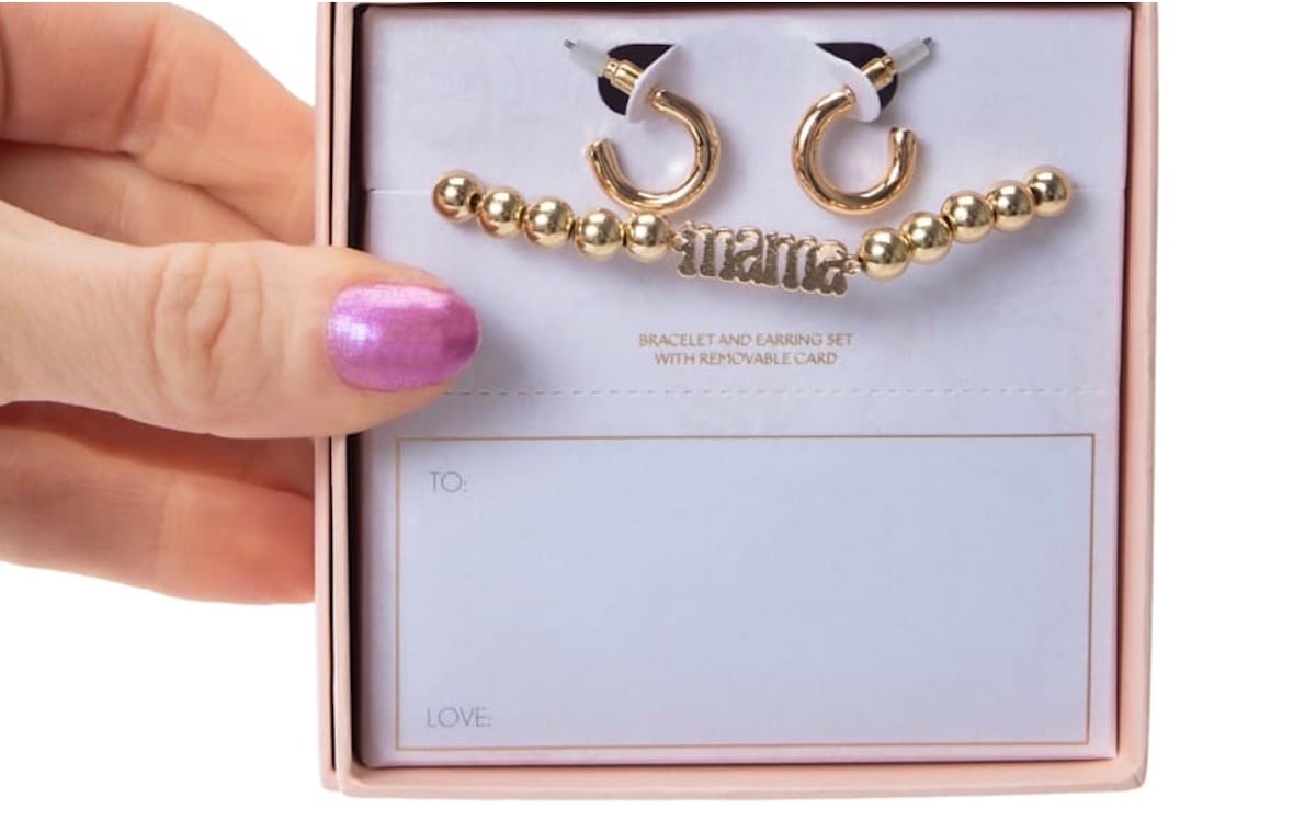 A hand with purple nail polish holds a five below gift box containing gold hoop earrings and a beaded "mama" bracelet. The box includes a card with "TO:" and "LOVE:" fields for easy gifting.