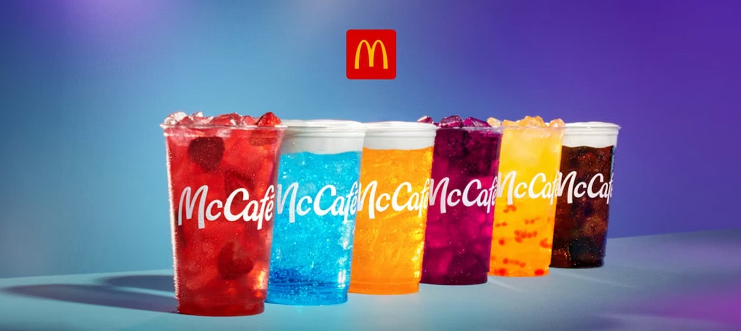 Six colorful McCafé drinks in clear cups, ranging from red, blue, orange, yellow, to dark purple, are lined up with ice, against a purple gradient background and a McDonald's logo above them.