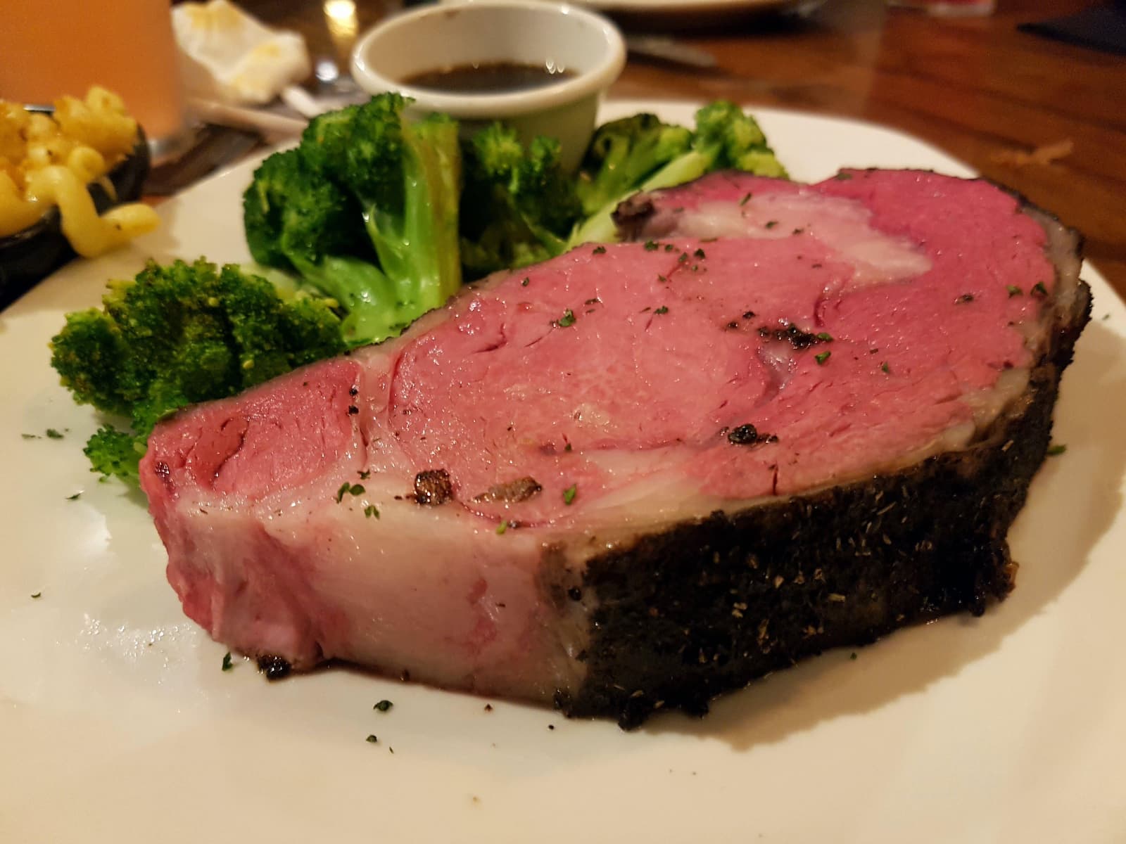 A thick slice of prime rib with a seasoned crust, served with steamed broccoli and a small cup of au jus on a white plate.