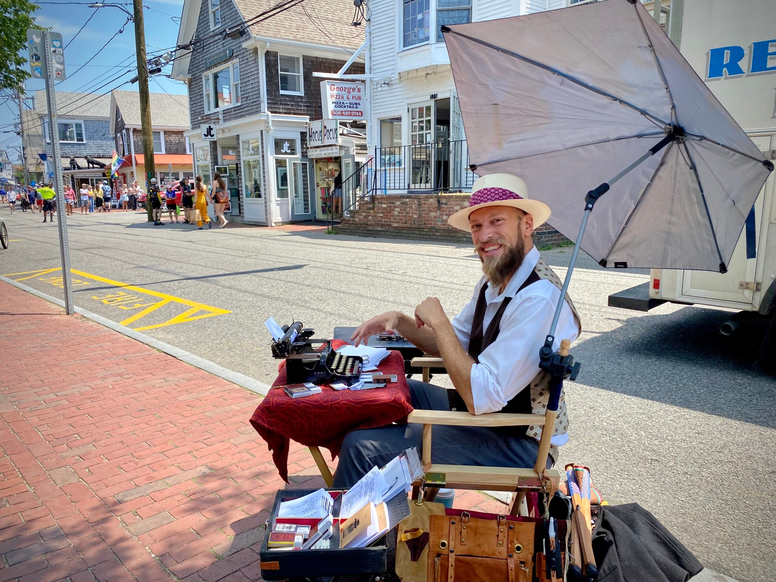 A man in vintage attire sits at a small outdoor desk with a typewriter on a sunny street, smiling at the camera. An umbrella shades him, and open mail and supplies surround his workspace. Shops and people are visible in the background.