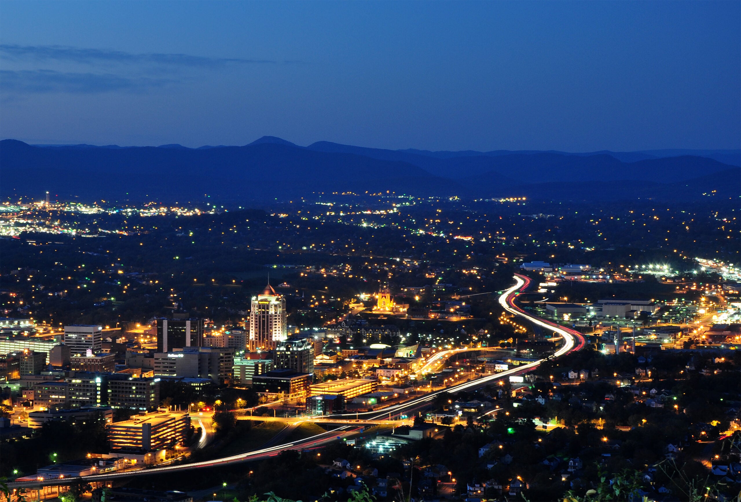 Cityscape at dusk with buildings lit up, a curving highway with light trails from cars, and mountains in the background under a darkening blue sky.