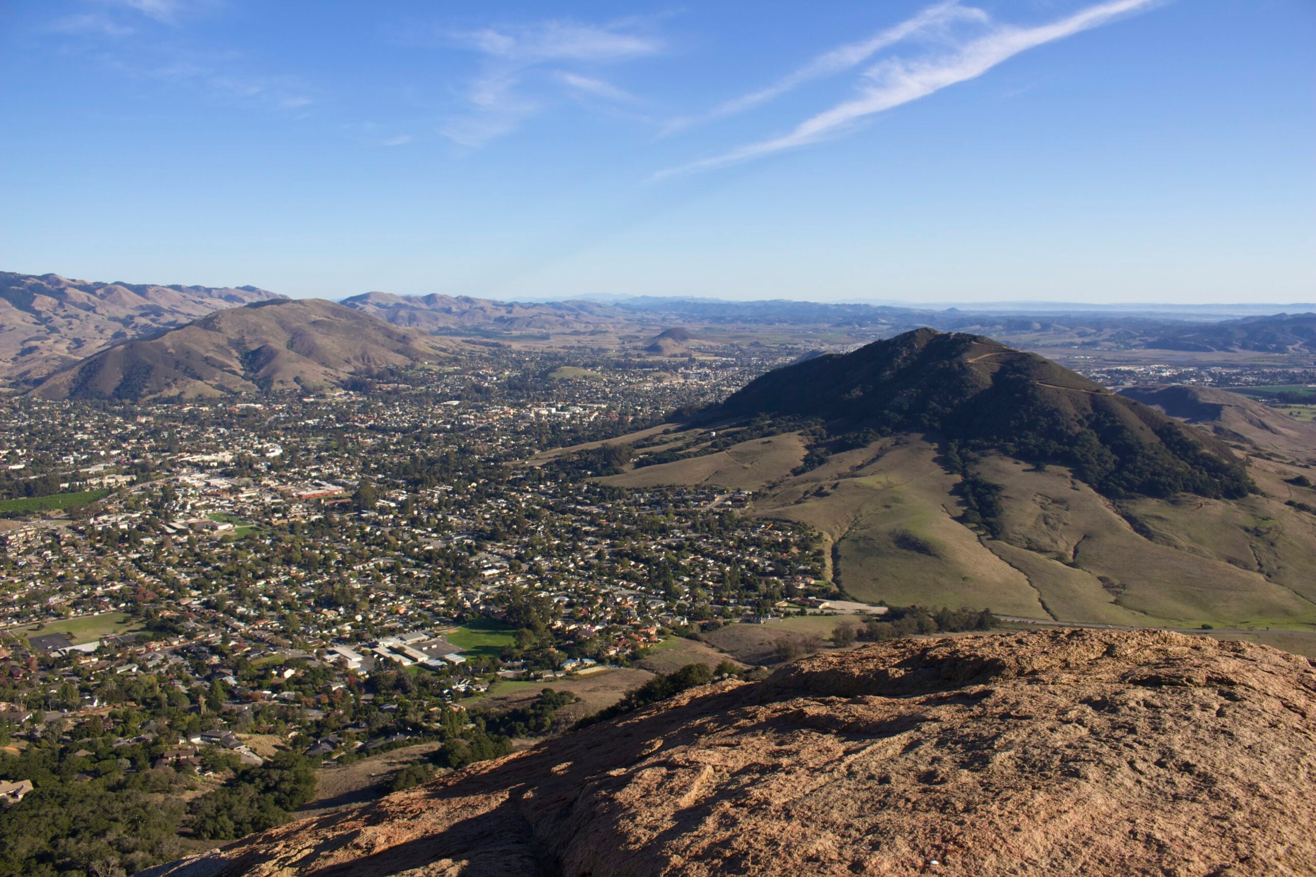 View from a rocky hilltop overlooking a valley with a small city, surrounded by green hills and mountains under a clear blue sky with a thin, wispy cloud.