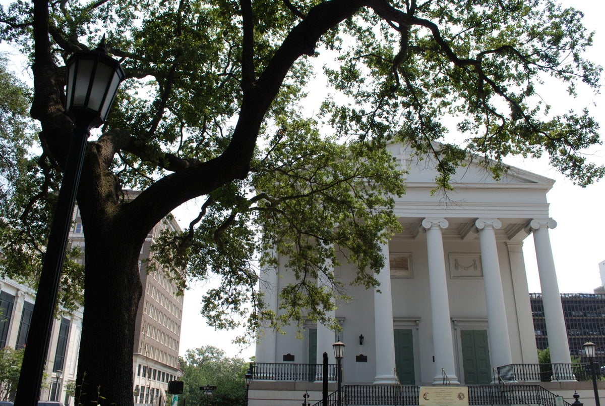 A large tree with sprawling branches stands in front of a white building with tall columns and green doors, partially obscuring its entrance. A black street lamp is also visible on the left.