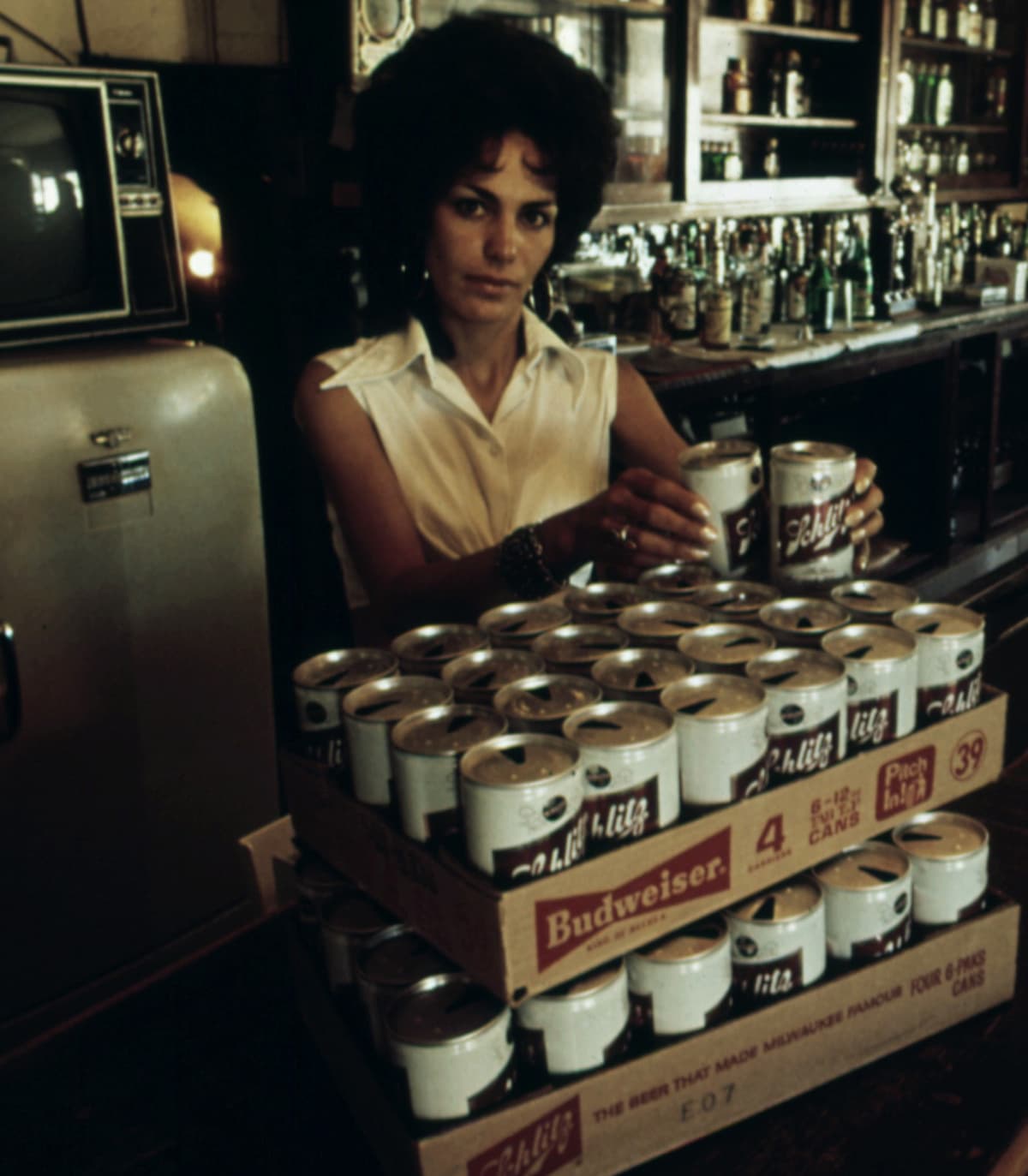 A woman with dark curly hair arranges stacks of Schlitz beer cans on a bar counter, with two cardboard trays labeled Budweiser in front of her. A vintage refrigerator and shelves of bottles are visible in the background.