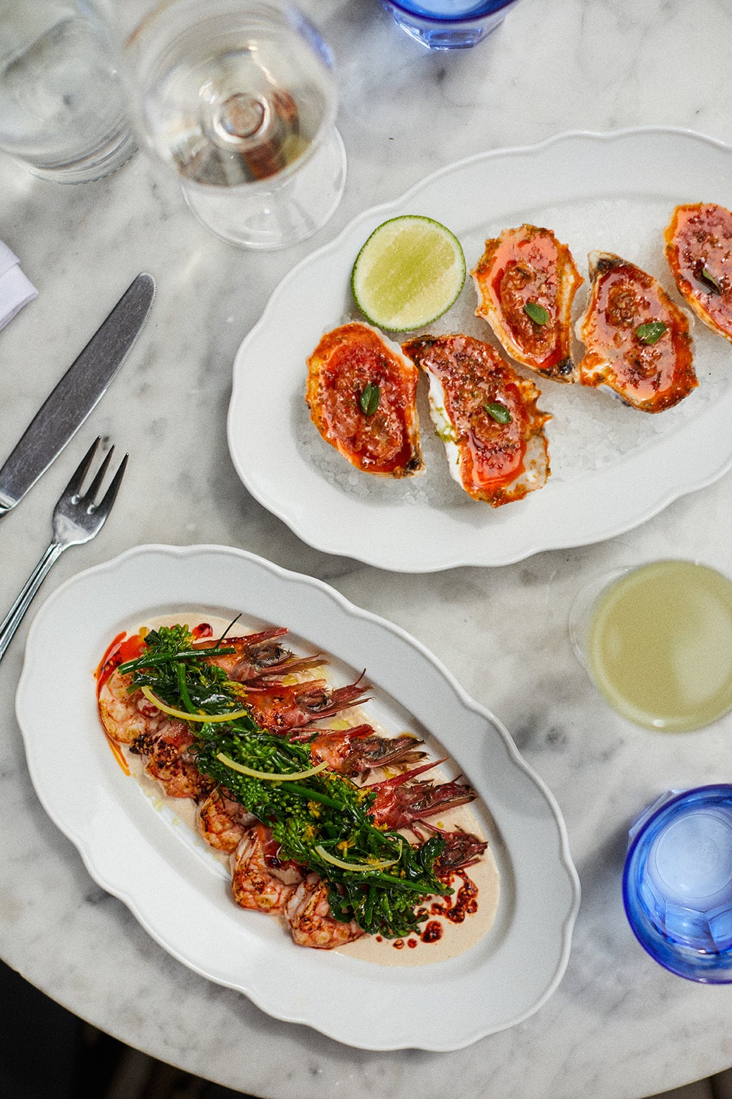 Two white plates on a marble table: one with oysters topped with red sauce and herbs, and another with sliced seafood, greens, and sauce. Drinks and blue water glasses are also visible.