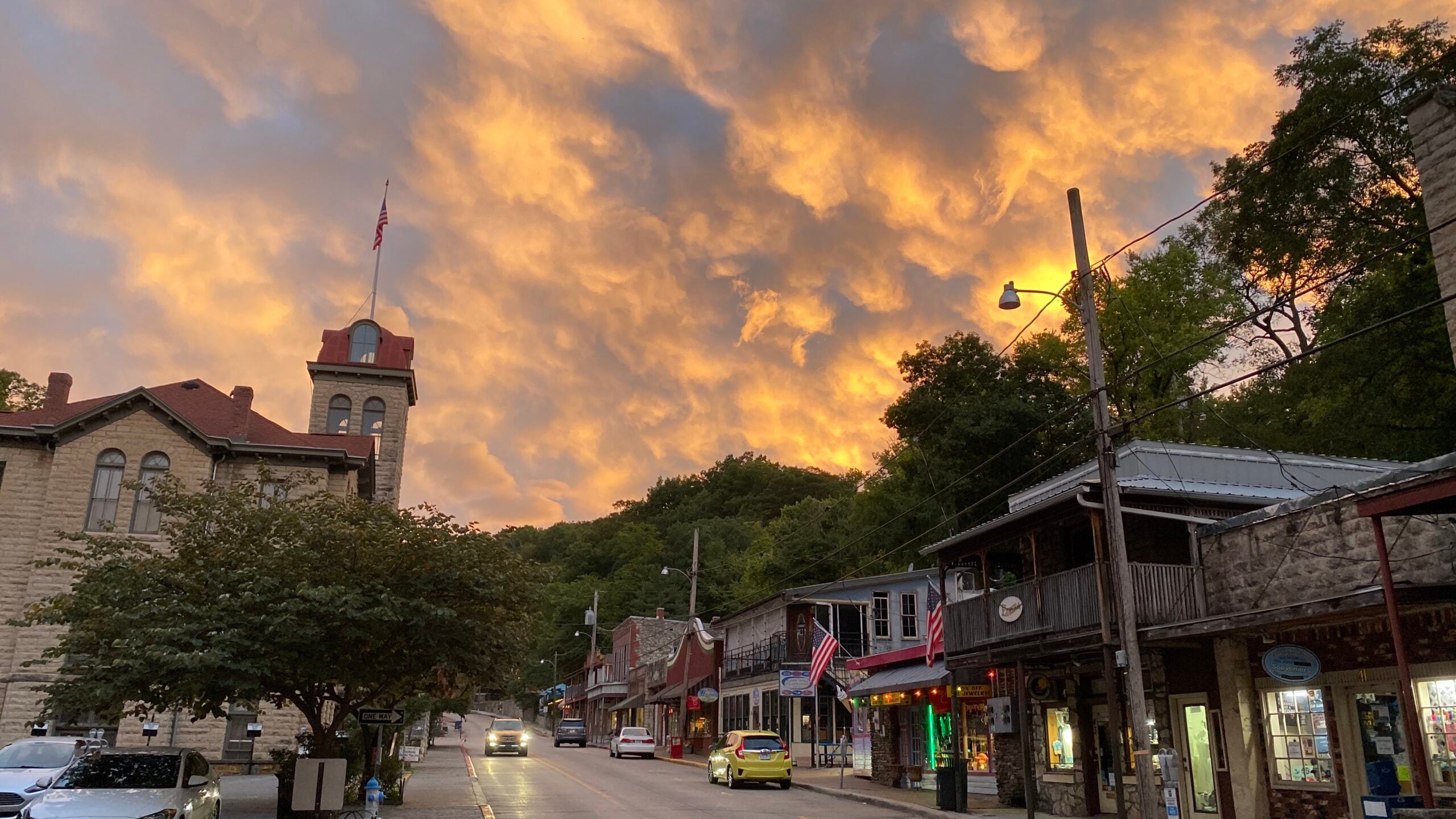 A small town street at sunset, with dramatic orange clouds in the sky, historic buildings line the road, some with American flags, cars parked and trees in the background.