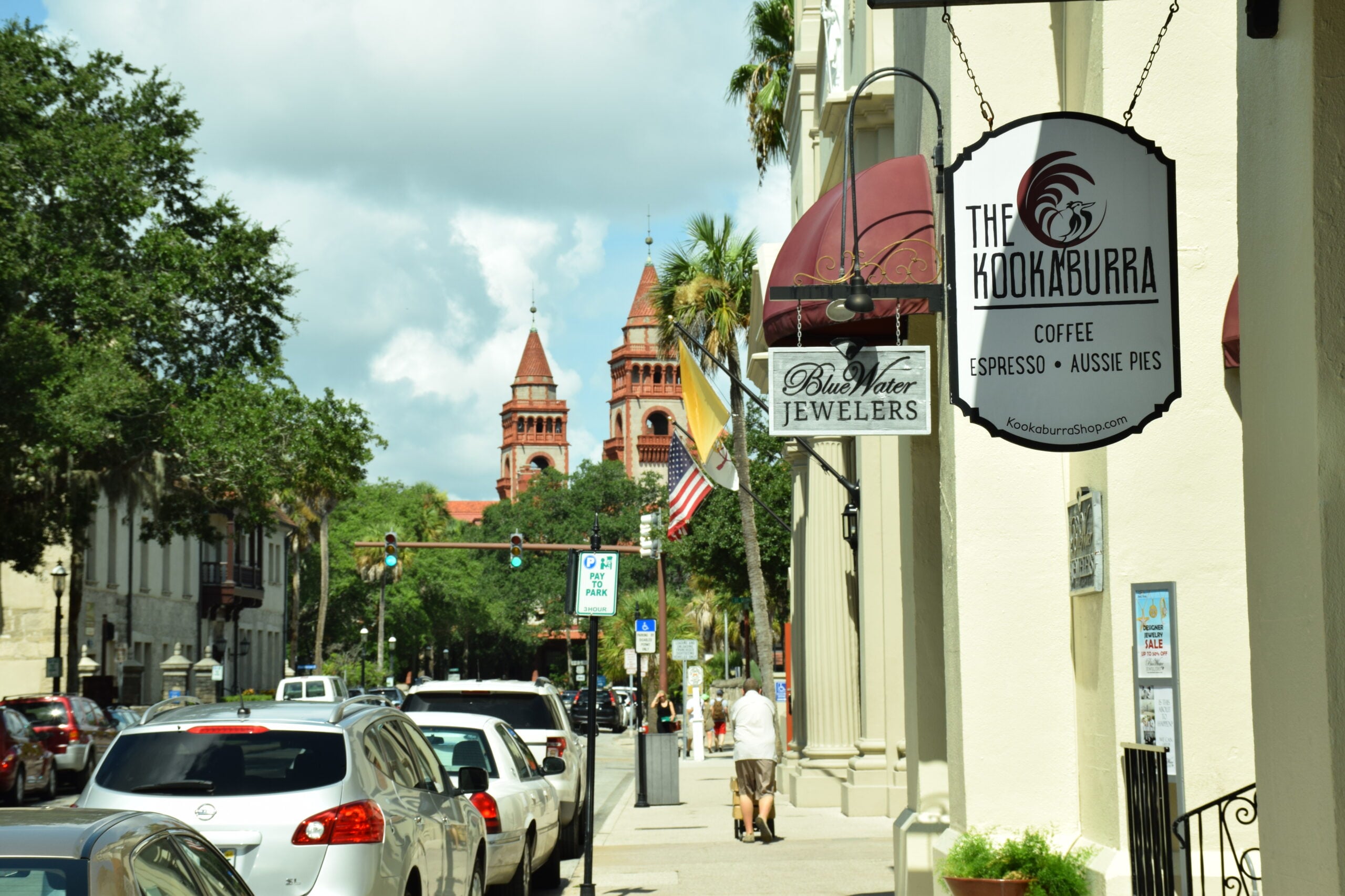 A sunny street lined with parked cars, shops, and palm trees, with a sign for "The Kookaburra Coffee" in the foreground and red-roofed towers visible in the background.