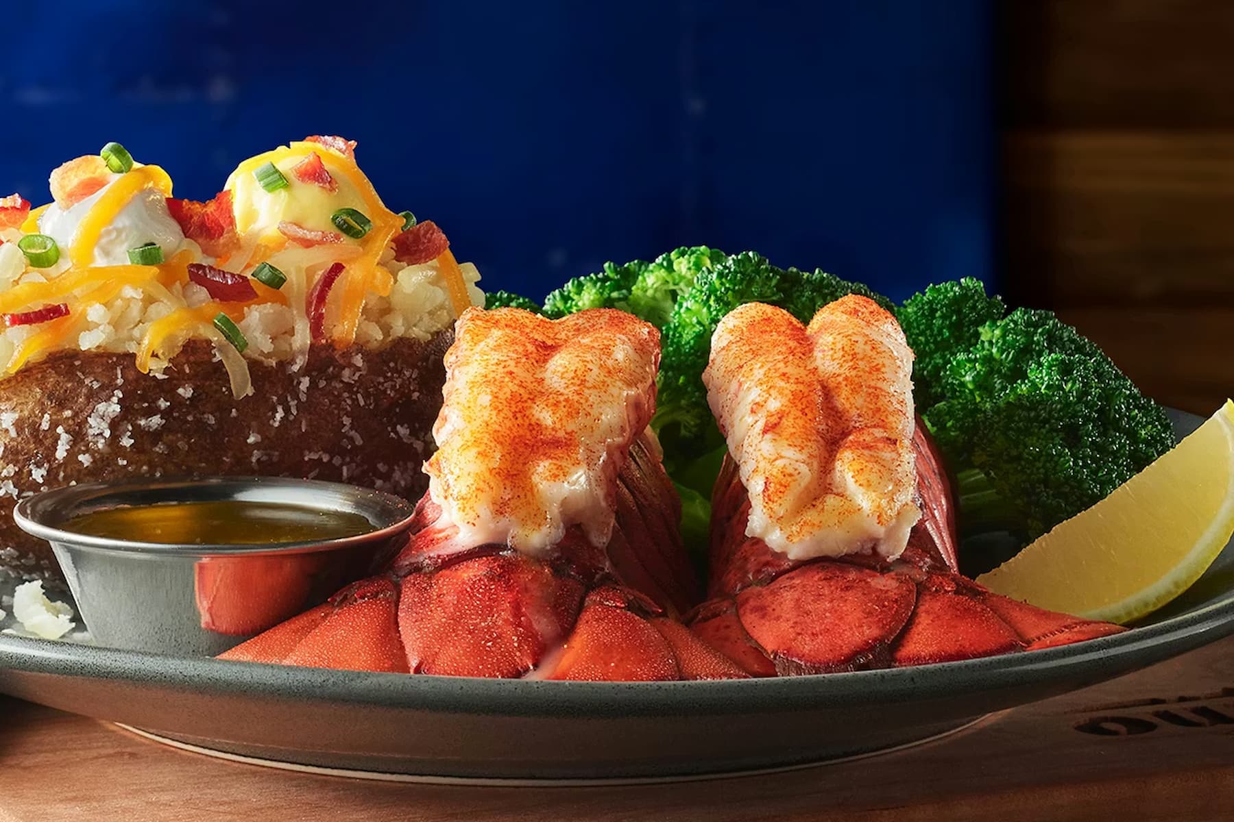 Two broiled lobster tails with melted butter, a loaded baked potato topped with cheese, bacon, and chives, steamed broccoli, and a lemon wedge on a plate.