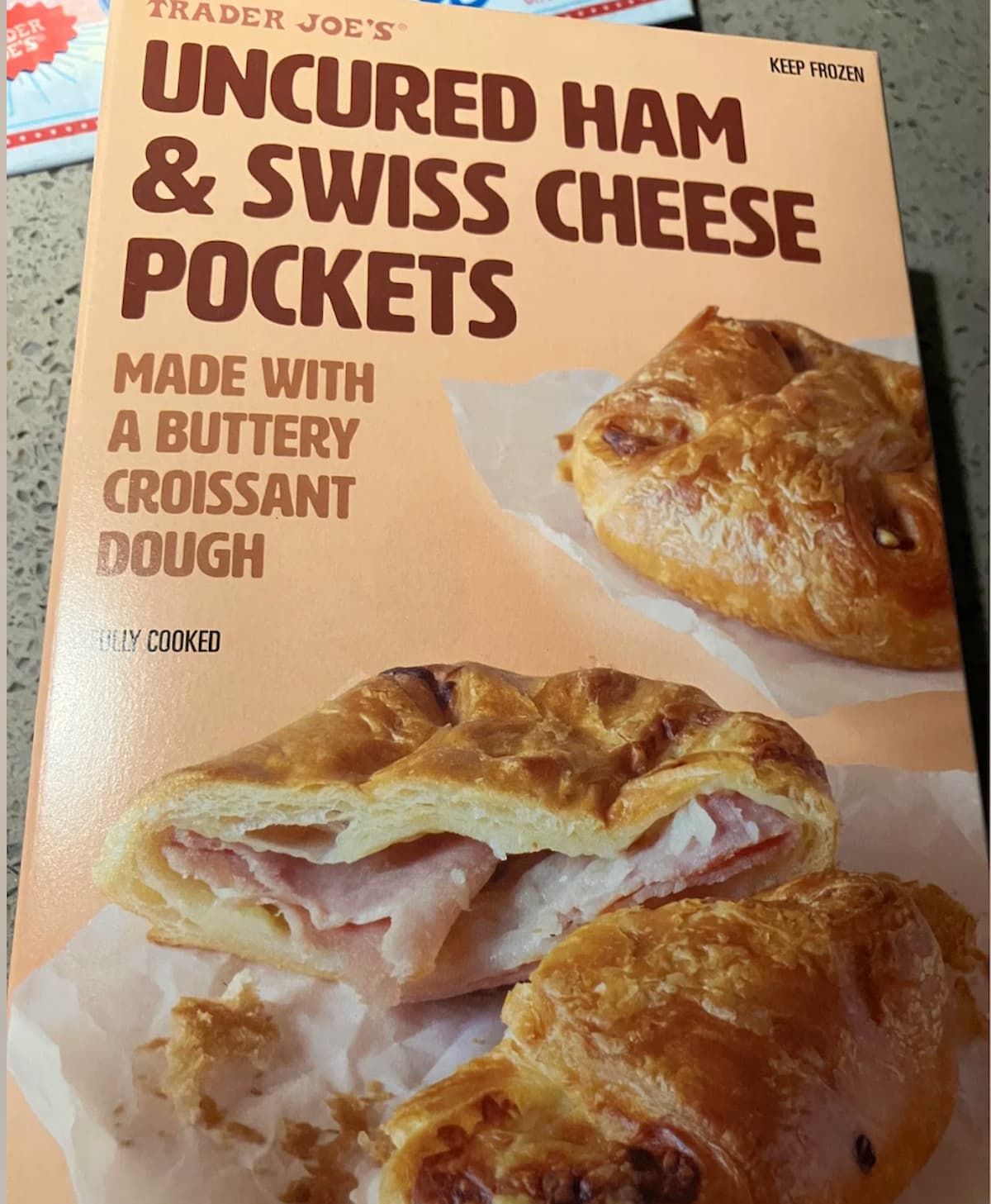 A box of Trader Joe's Uncured Ham & Swiss Cheese Pockets, featuring an image of flaky croissant dough pockets filled with ham and cheese on the front, with bold product text and serving suggestion.