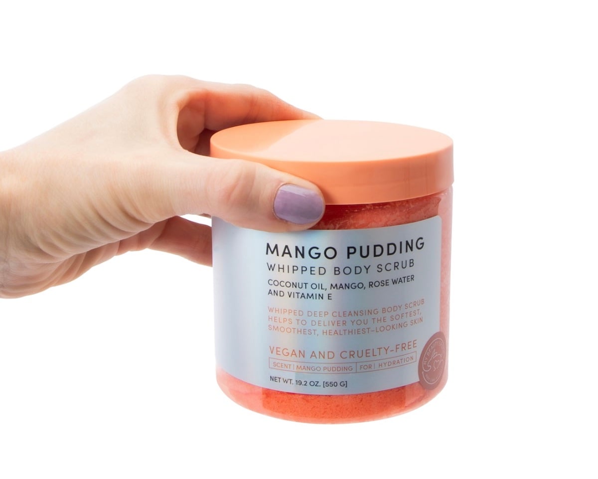 A hand with lavender nail polish holds a jar of Mango Pudding Whipped Body Scrub with a peach lid. The label highlights vegan, cruelty-free ingredients like coconut oil, mango, rose water, and vitamin E—available now at Five Below.