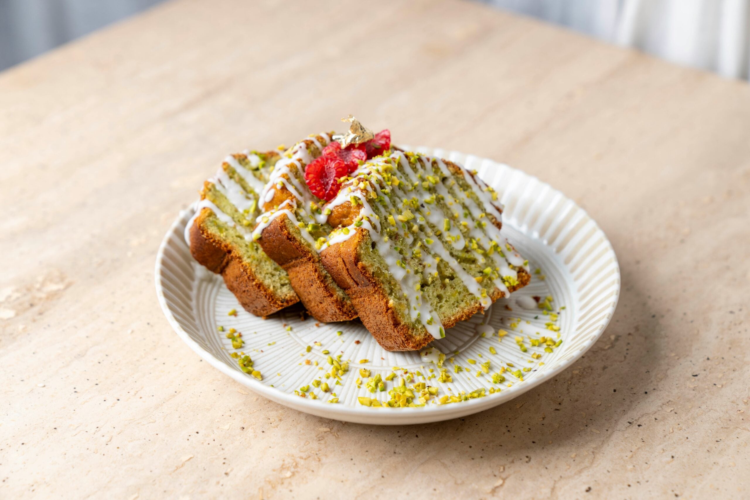 Slices of pistachio cake topped with white icing, crushed pistachios, and raspberries are arranged on a white plate set on a light-colored marble surface.