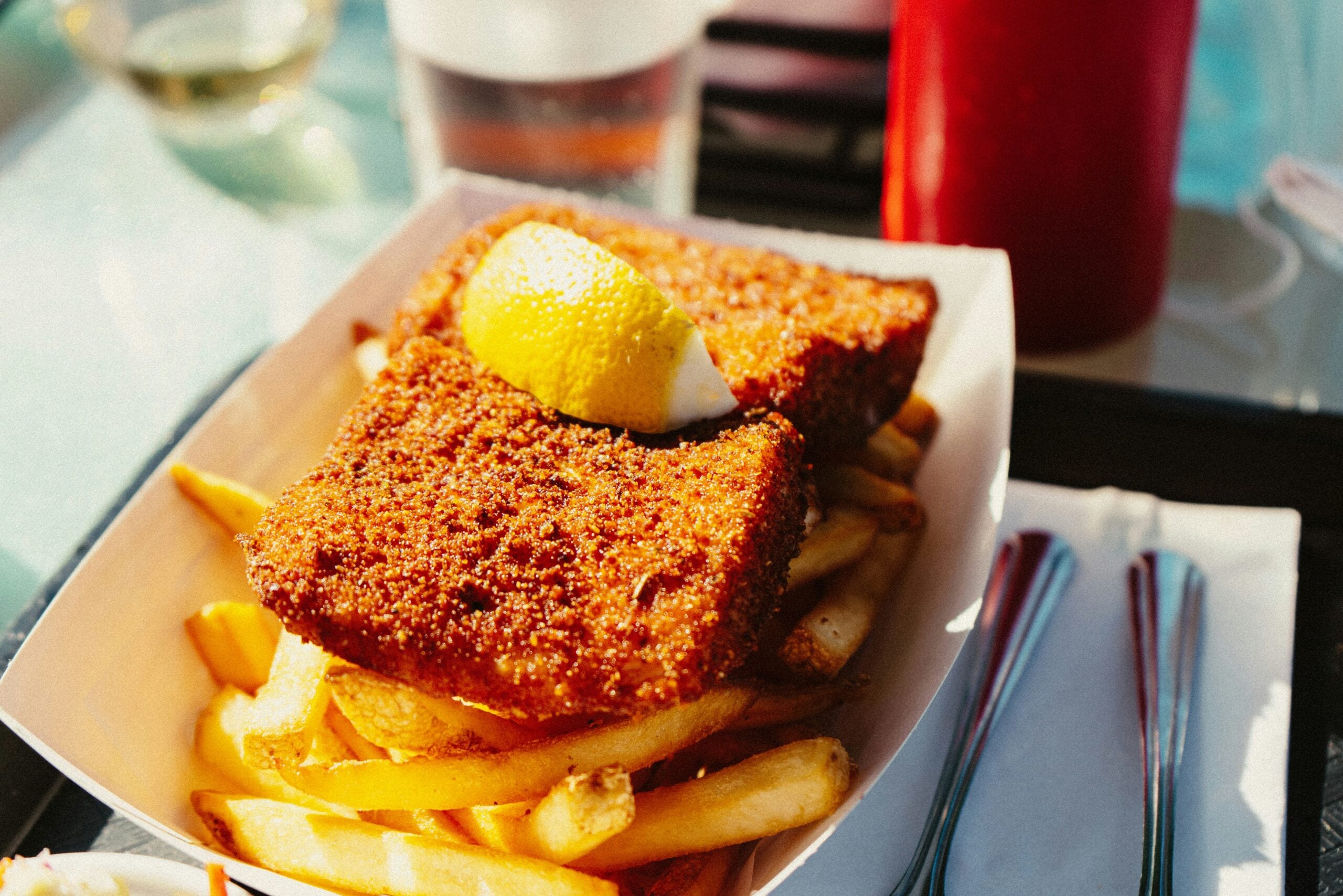 A serving of golden French fries topped with two breaded, fried fish fillets and a lemon wedge, in a paper tray next to a fork and knife. A glass of water and a ketchup bottle are in the background.