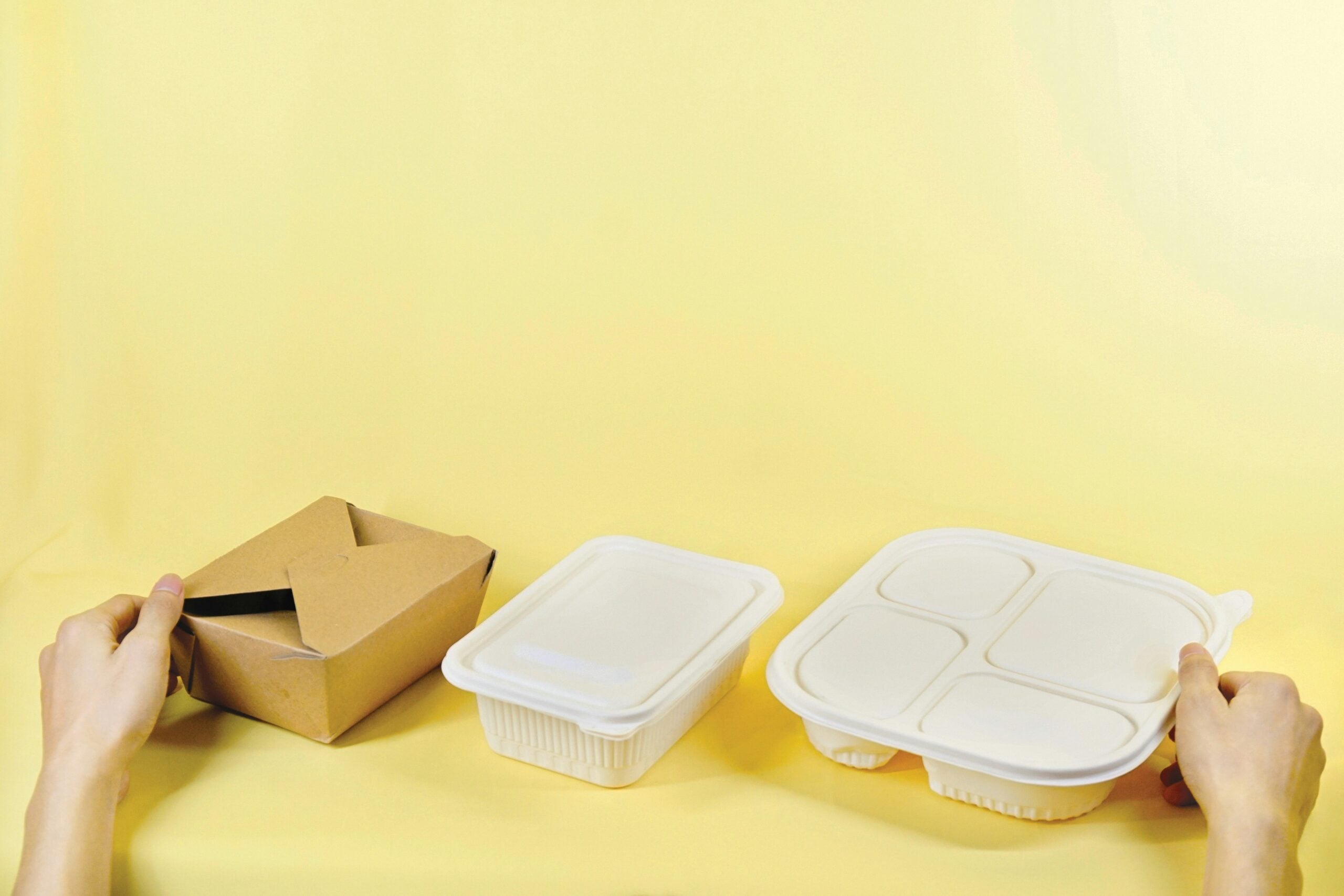 Two hands hold different types of food containers against a yellow background: a brown paper box on the left, a rectangular plastic container in the center, and a white plastic compartment tray on the right.
