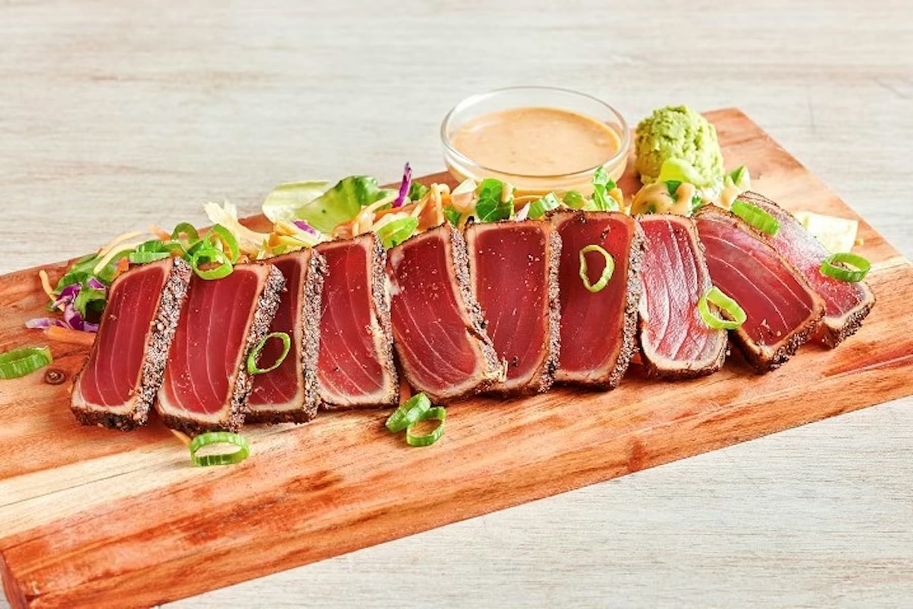 Slices of seared ahi tuna topped with green onions are arranged on a wooden board, accompanied by salad, a dollop of wasabi, and a small dish of dipping sauce.