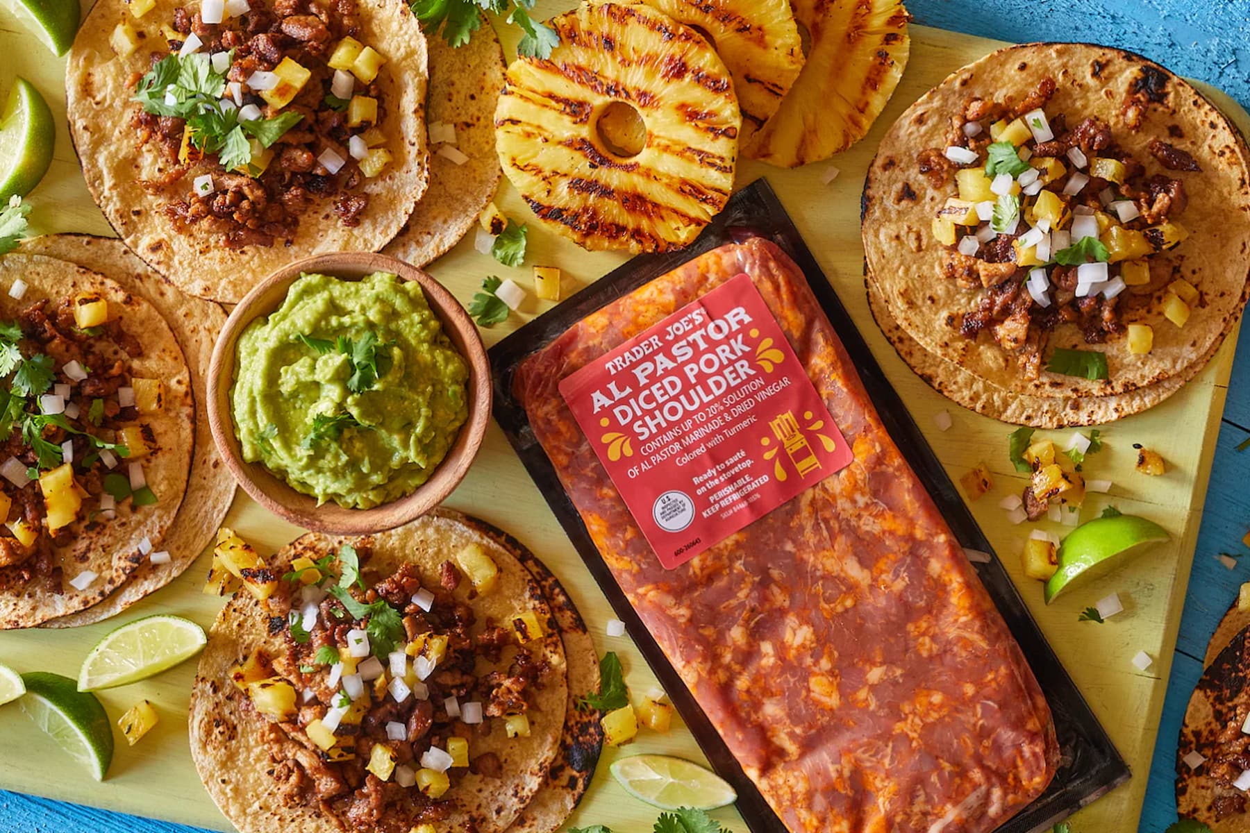 Five tacos filled with seasoned pork, diced onions, cilantro, and pineapple sit on a board alongside lime wedges, grilled pineapple slices, a bowl of guacamole, and a packaged tray of Trader Joe’s Al Pastor diced pork shoulder.