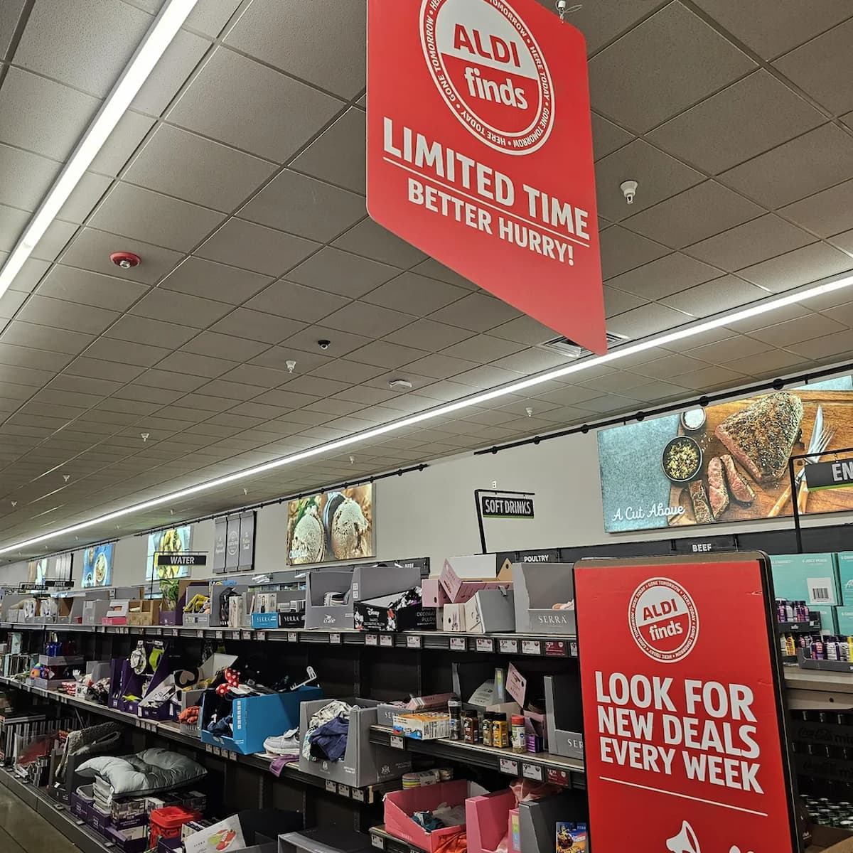 Aldi store aisle with shelves stocked with various products and a large red sign overhead reading "ALDI Finds, Limited Time, Better Hurry!" Above shelves are wall graphics of food and kitchen items.