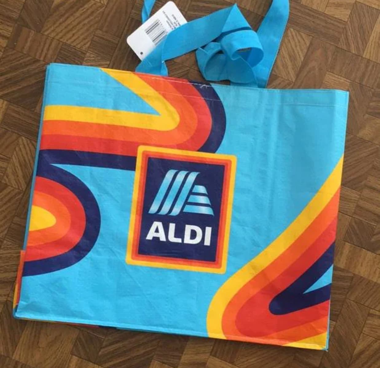 A reusable shopping bag with a blue background, retro orange and yellow swirls, and the ALDI logo in the center, lying flat on a wooden floor.