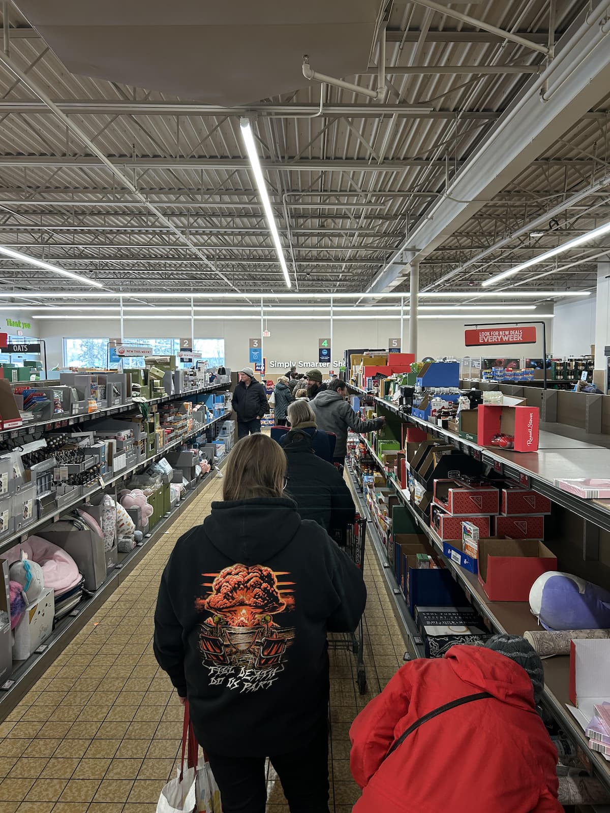 Shoppers stand in a single-file line with carts inside a store, surrounded by shelves stocked with various household goods and gift boxes. The store has a high ceiling with visible beams and fluorescent lights.