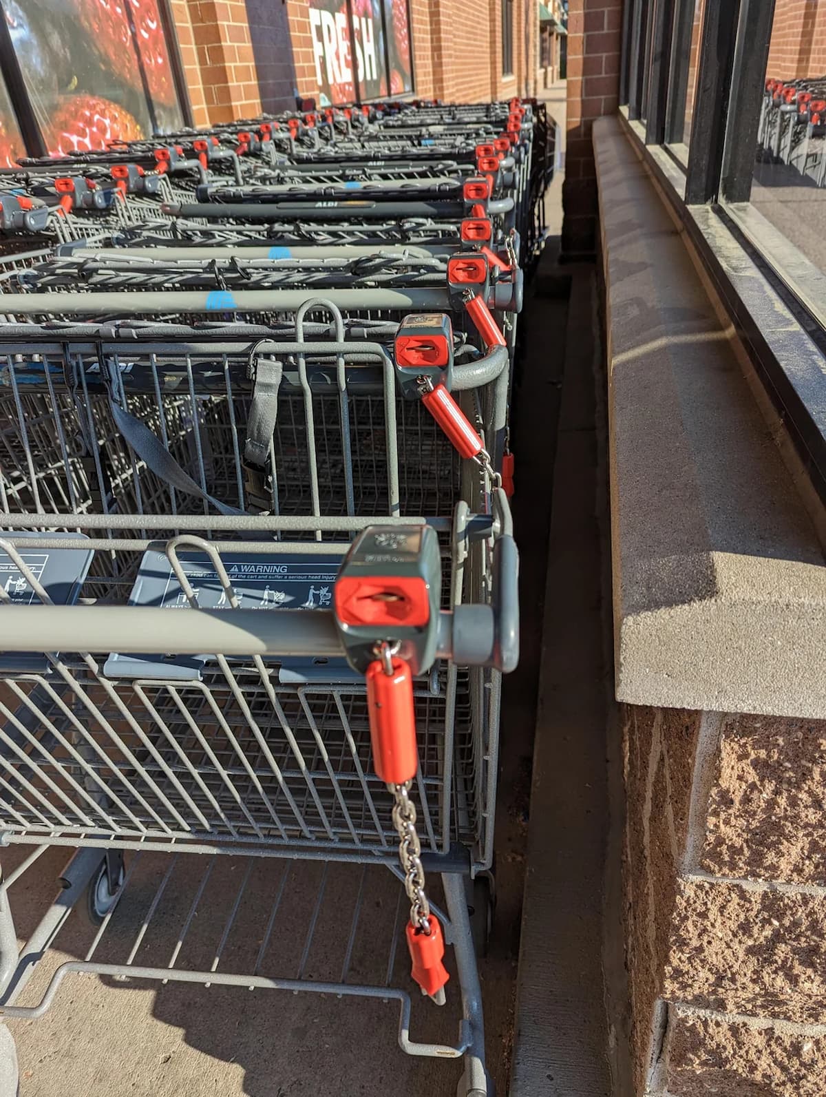 A row of shopping carts outside a store, each with a locking mechanism requiring a coin or token to release, lined up next to a brick wall in bright sunlight.