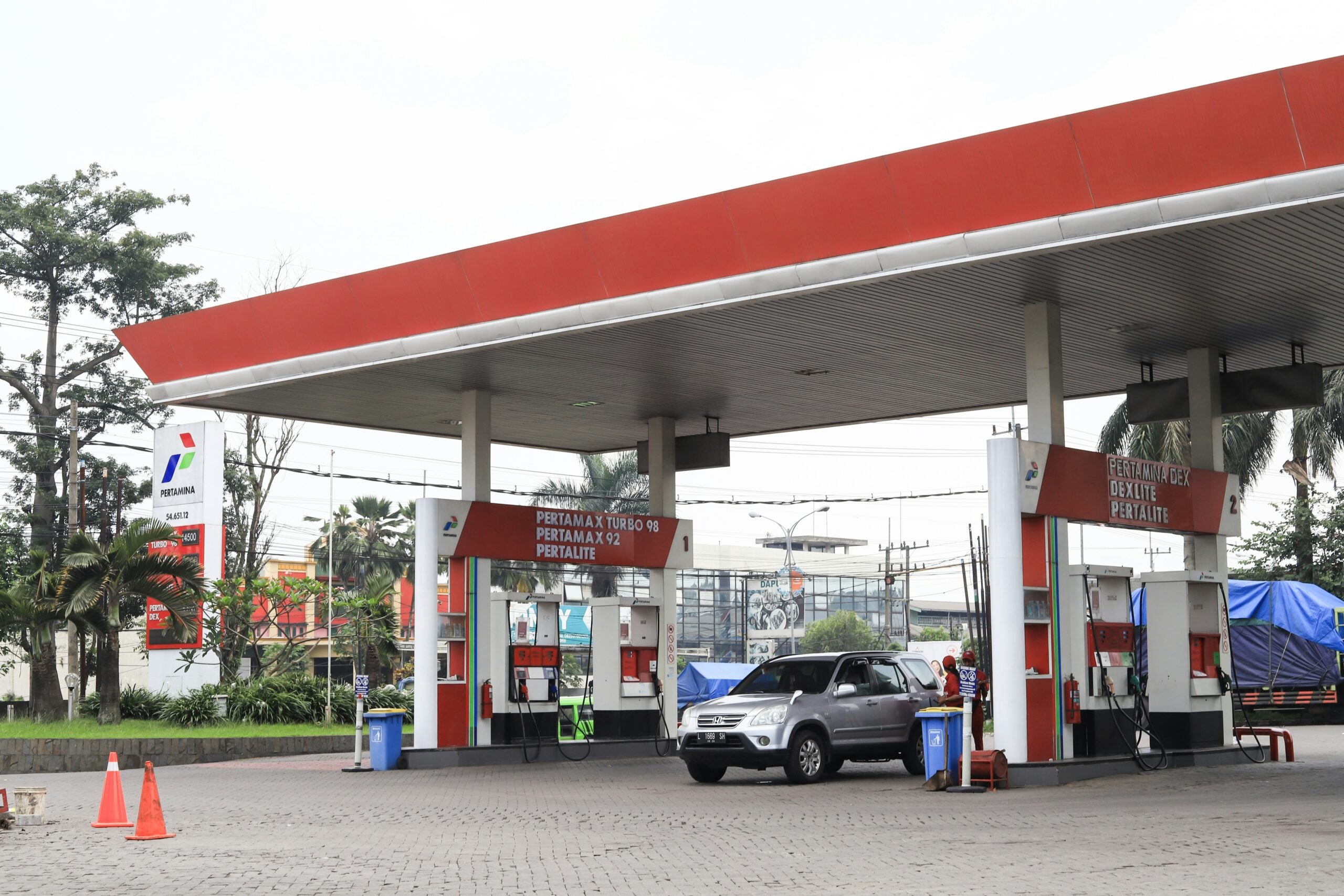 A silver SUV is parked at a gas station pump under a red and white canopy. The station has "PERTAMAX" and "PERTALITE" signs, with trees and other vehicles visible in the background.