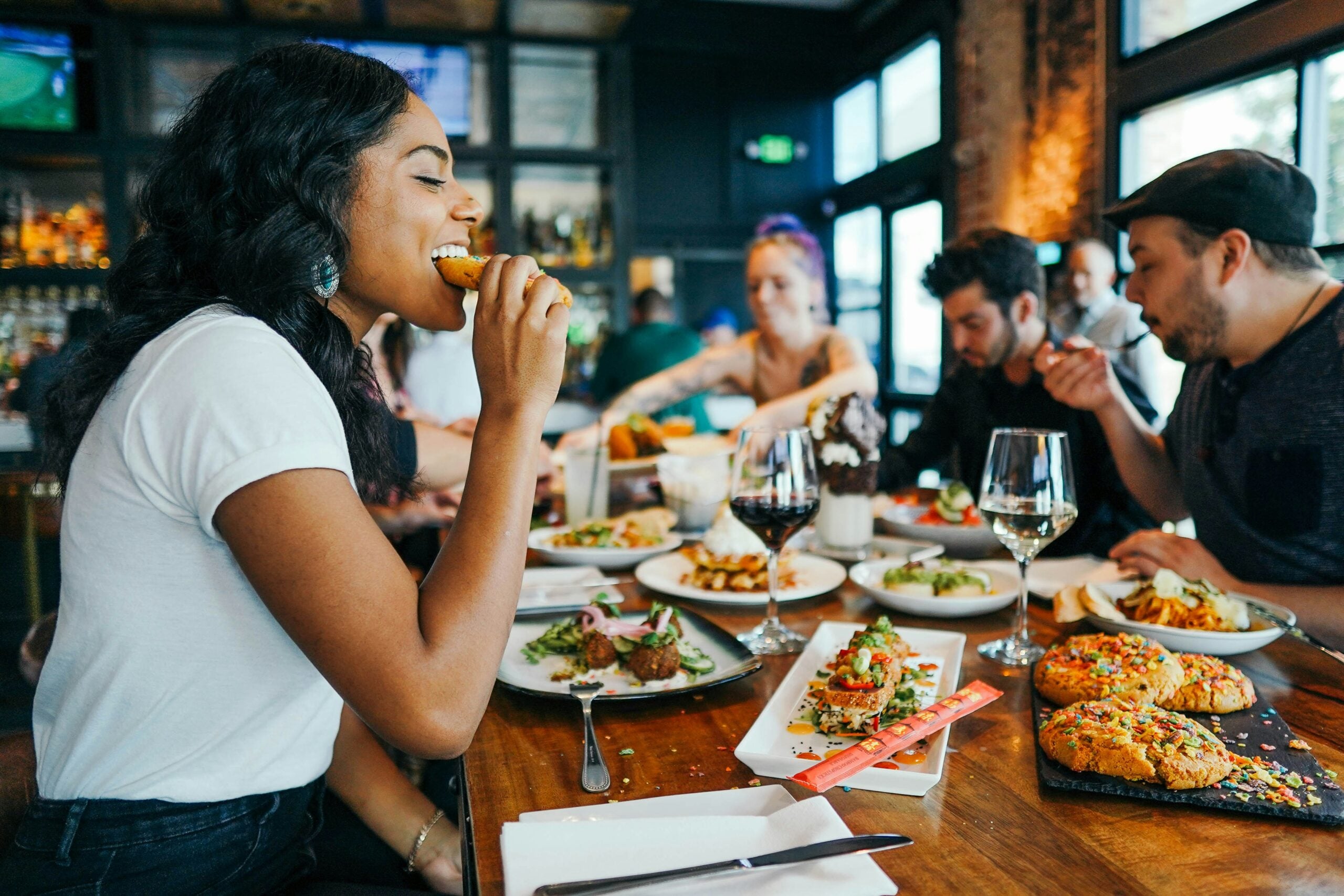 A group of people sit at a restaurant table enjoying a meal together. A woman in the foreground is smiling while eating, and various dishes and drinks are spread across the table. The setting appears lively and social.