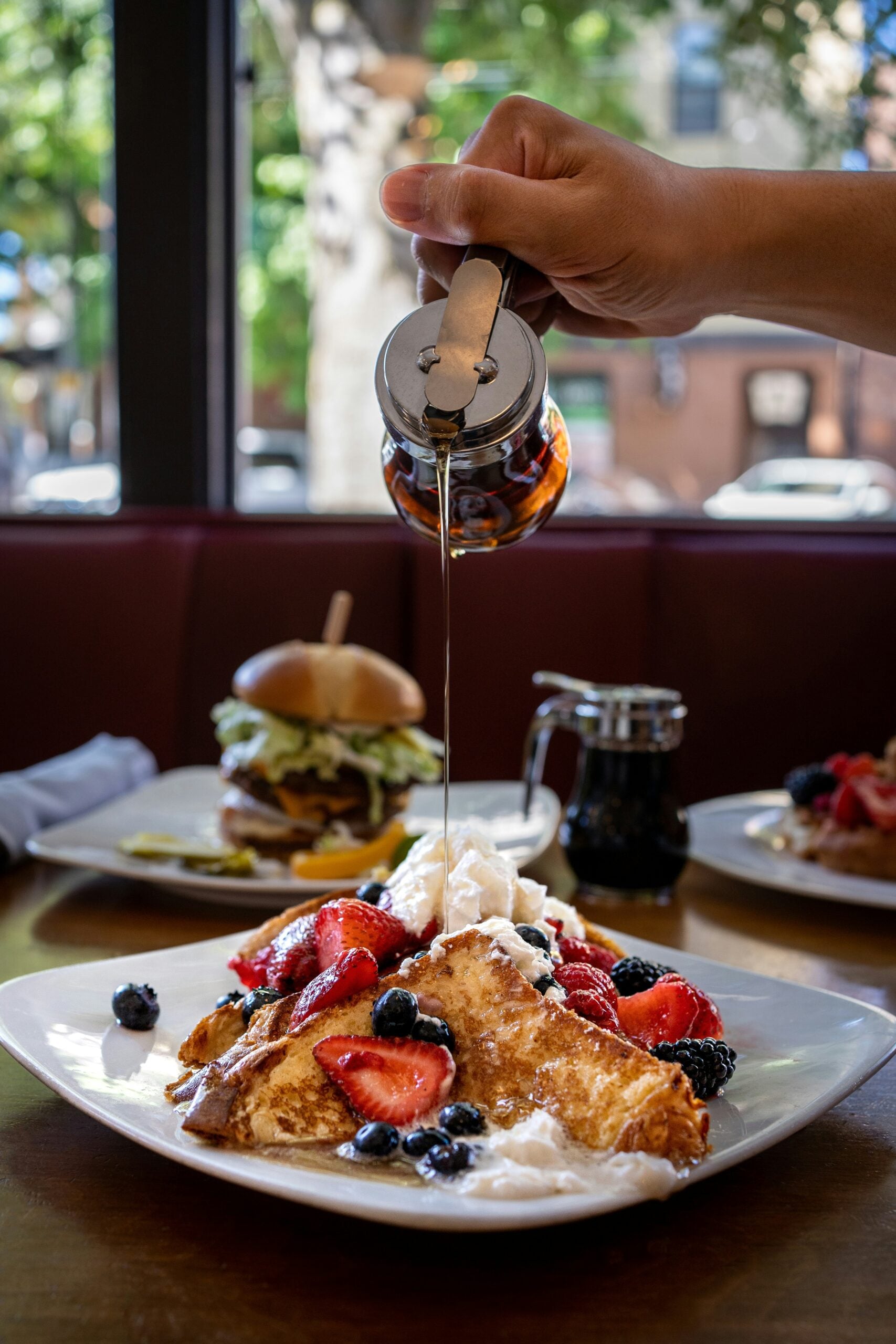 A hand pours syrup onto crepes topped with whipped cream, strawberries, blackberries, and blueberries. In the background, a plate with a burger and another with fruit-topped pancakes are visible.