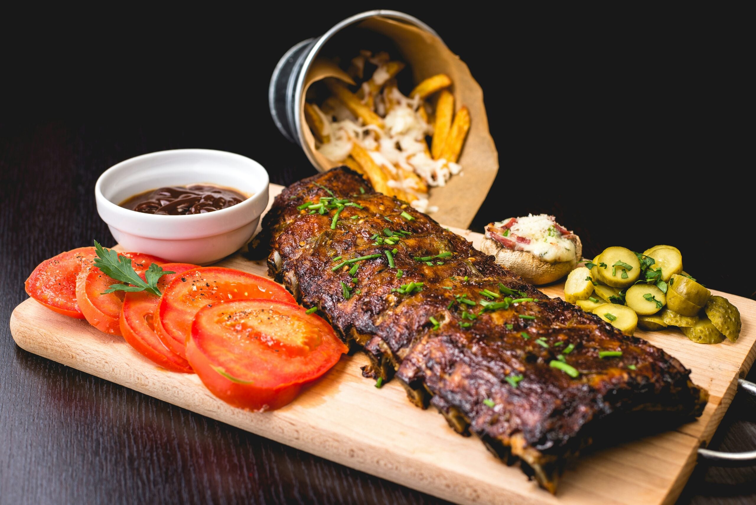 A wooden board with barbecued ribs garnished with herbs, sliced tomatoes, pickles, a baked potato with cream, a cup of barbecue sauce, and a small bucket of fries with coleslaw on a dark table.