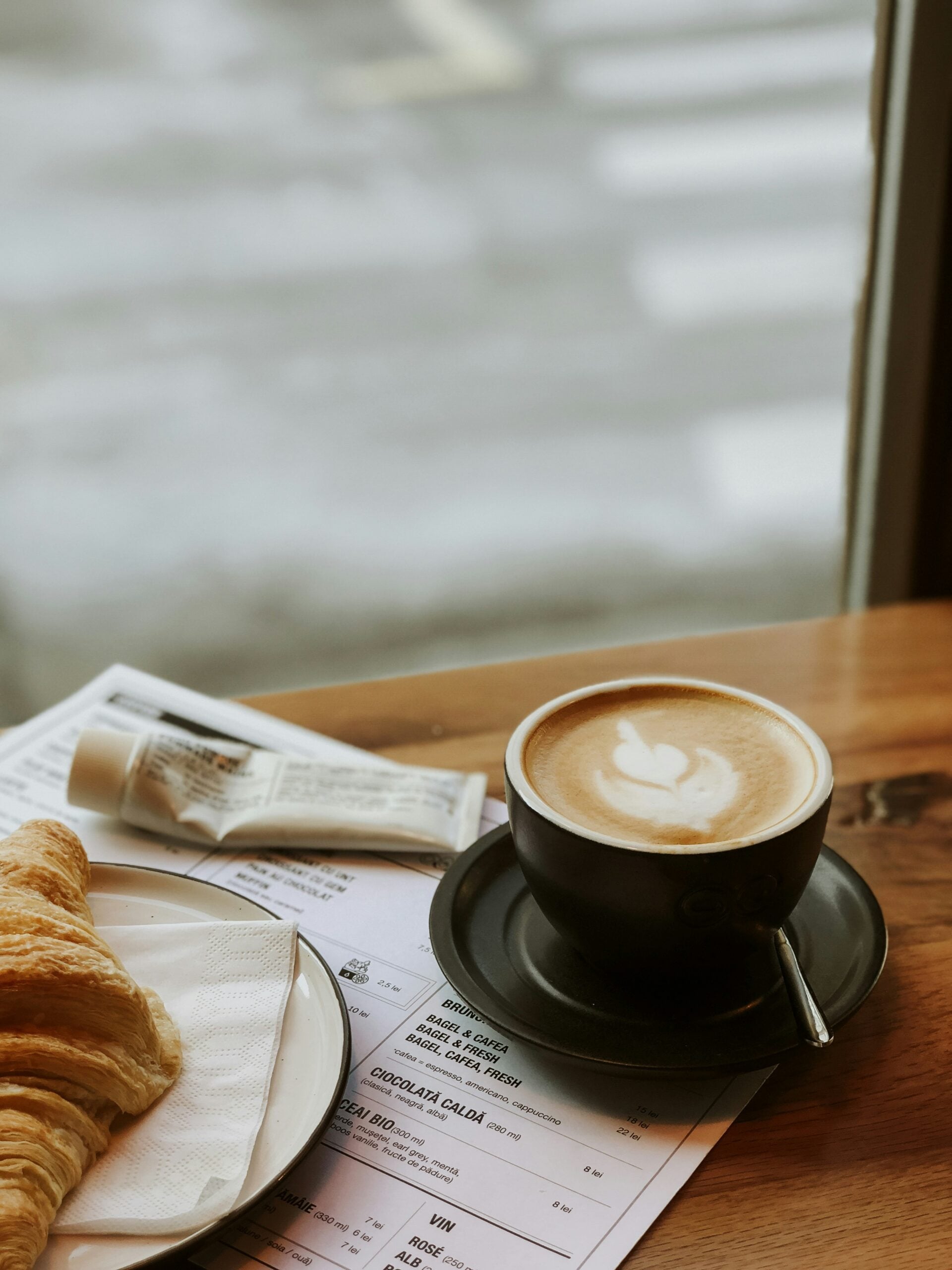 A cappuccino with latte art in a dark cup and saucer sits on a wooden table next to a croissant on a white plate, a tube of cream, and some printed receipts, near a window with a blurred street view outside.